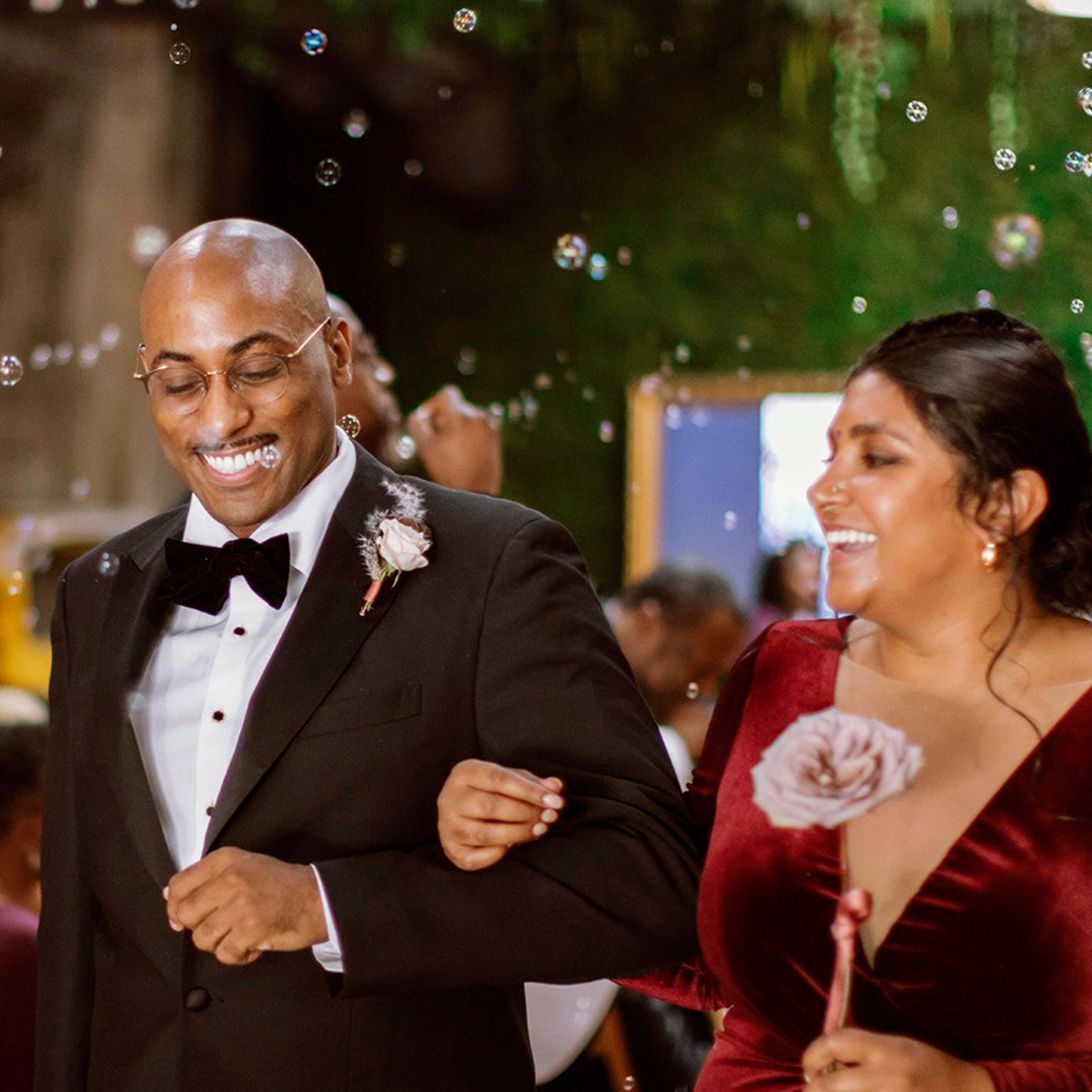 A man in a tuxedo and a woman in a red dress walk together among guests celebrating with bubbles at a wedding.