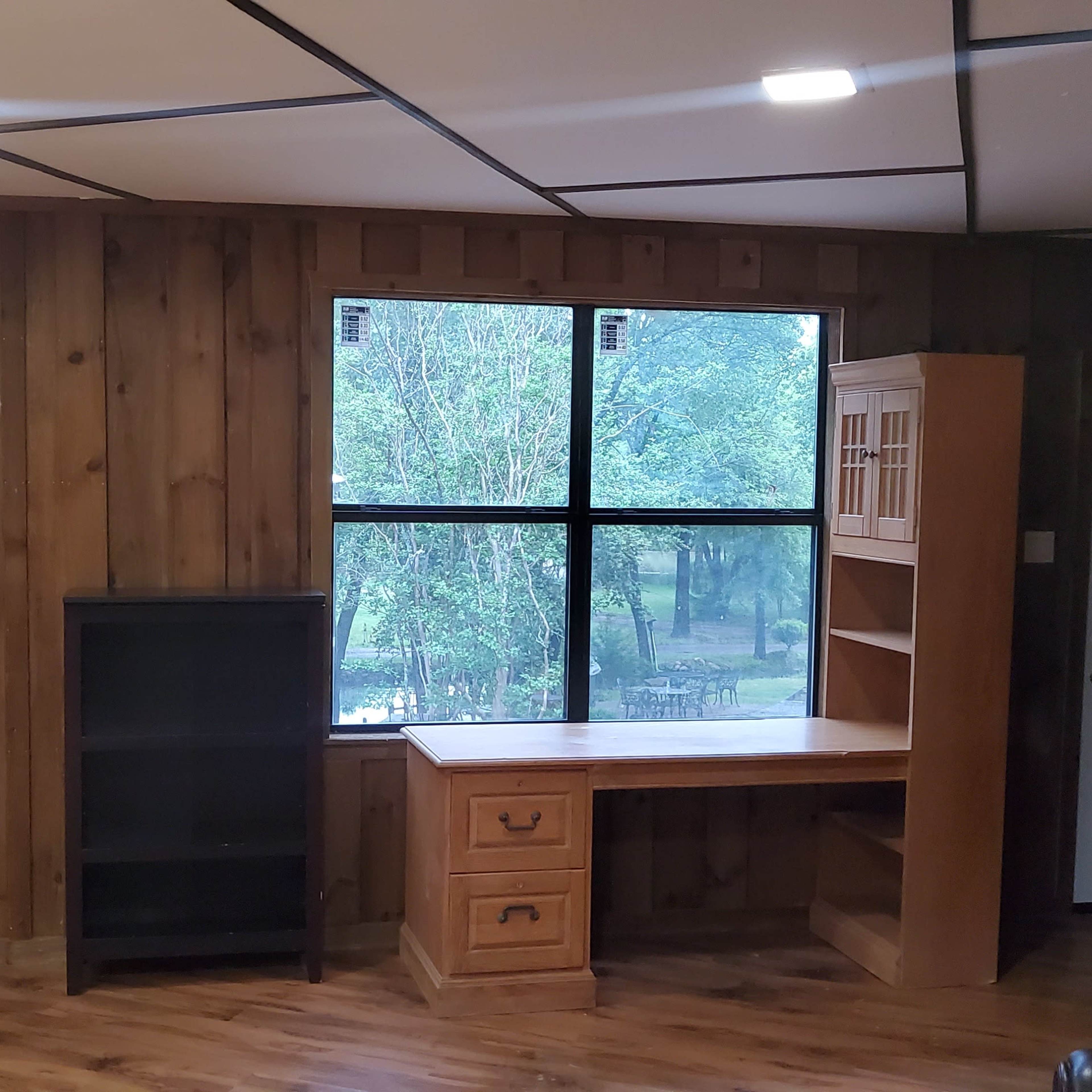 The room features wooden paneling, a large window overlooking a wooded area, a desk with drawers, a bookshelf, and a door leading outside.
