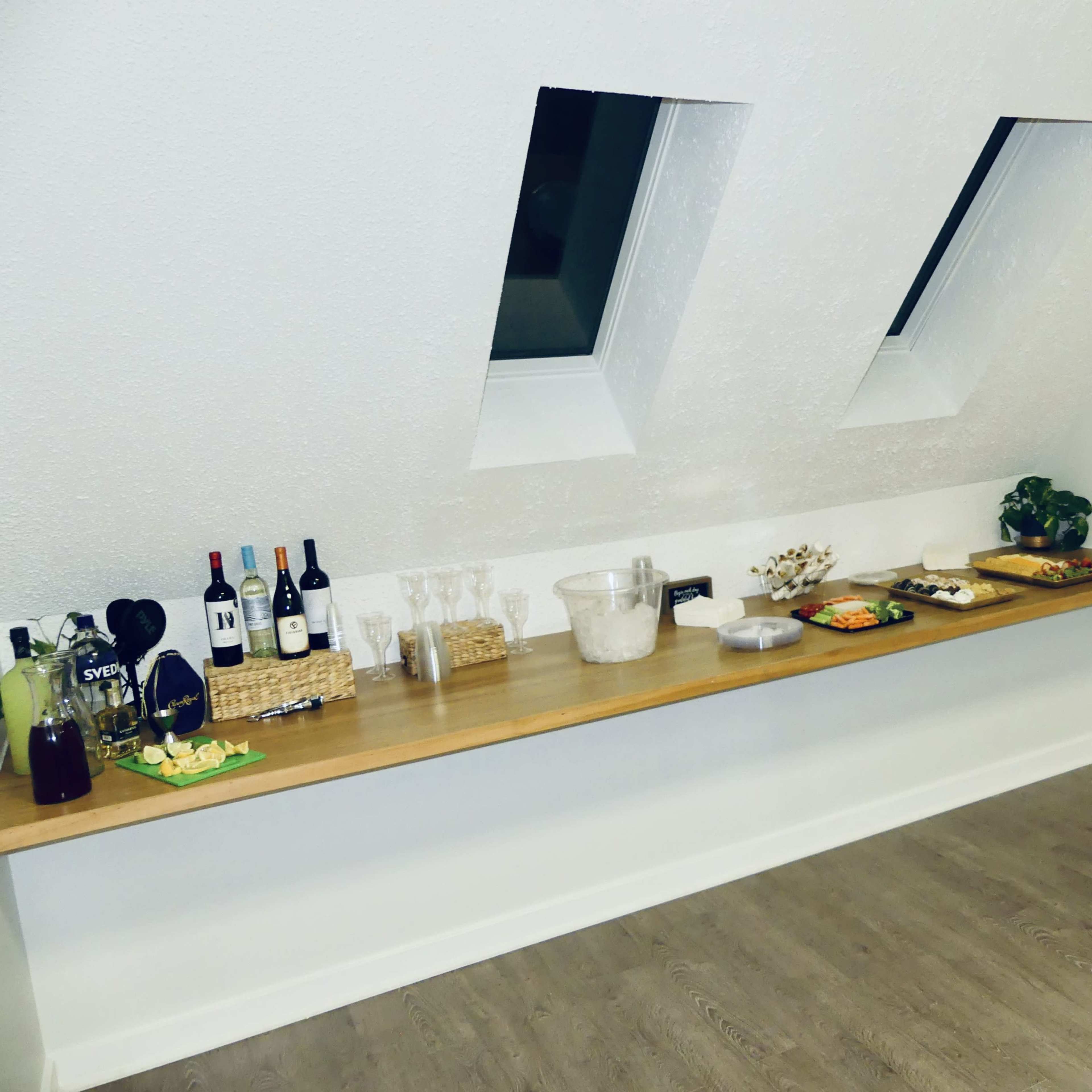 A wooden shelf displays bottles of wine, glasses, and various food items against a white wall with angled windows.