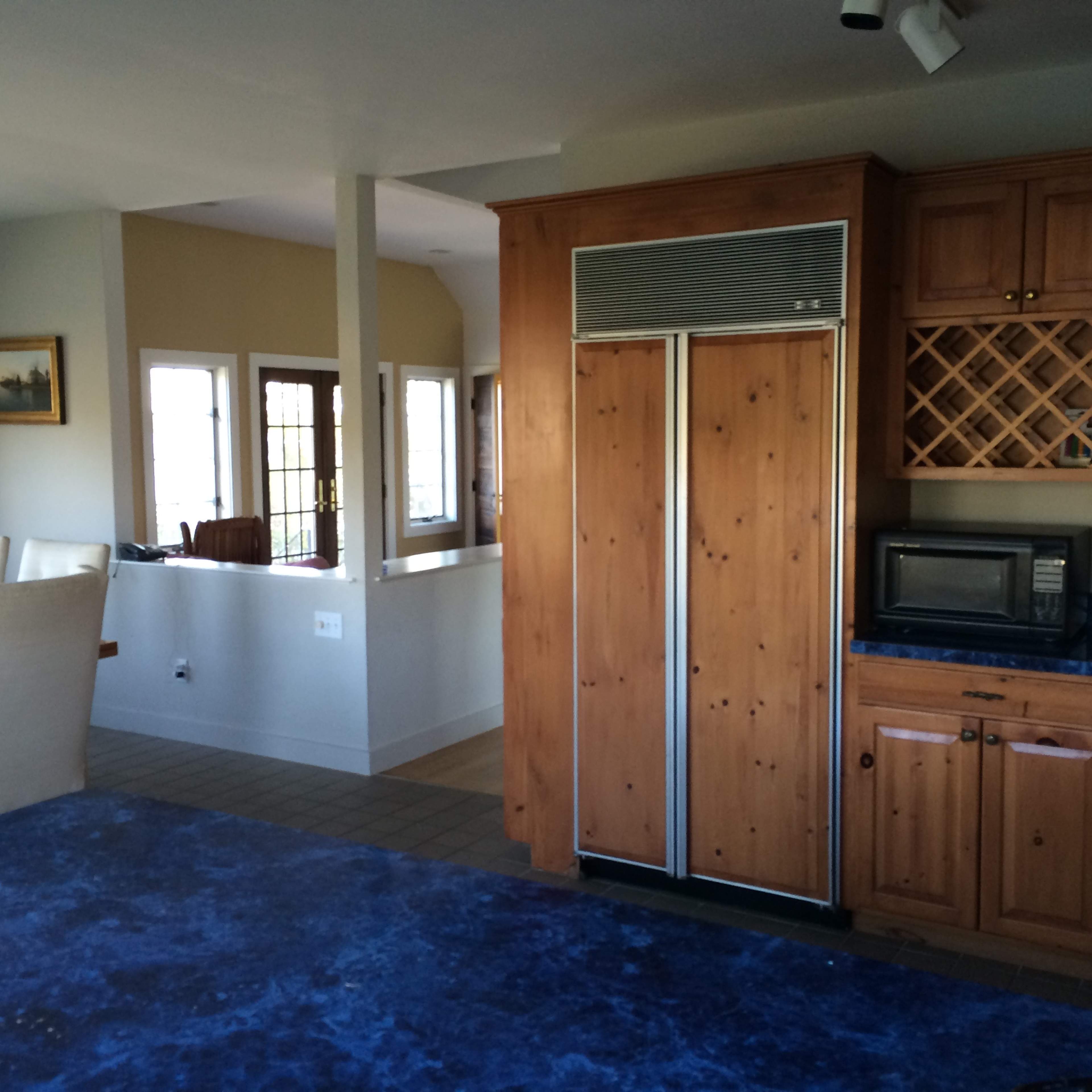 A kitchen with wooden cabinetry, a large stainless steel refrigerator, and a blue countertop, featuring a dining area and windows in the background.
