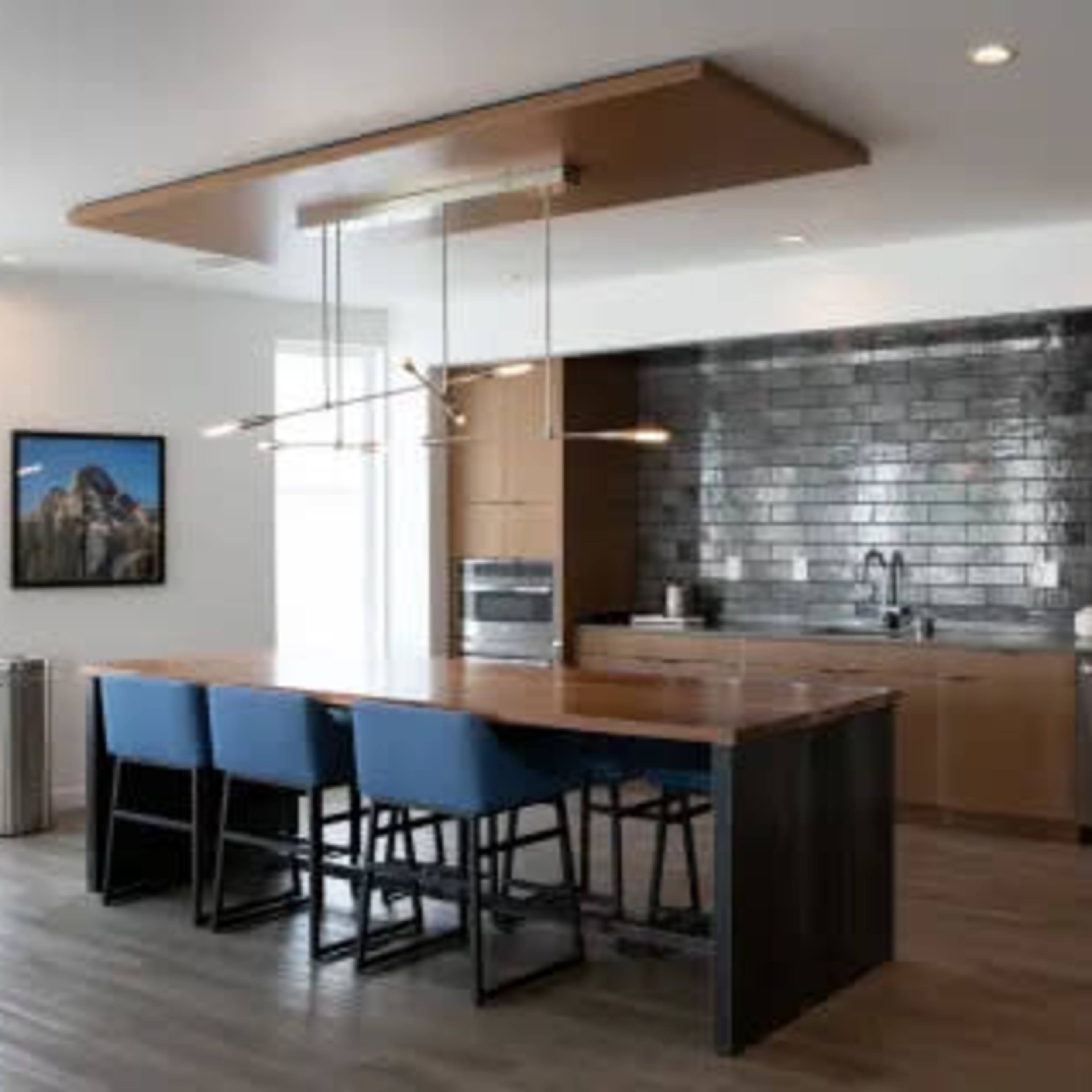 A modern kitchen features a large island with blue chairs, stainless steel appliances, and dark tiled backsplash.