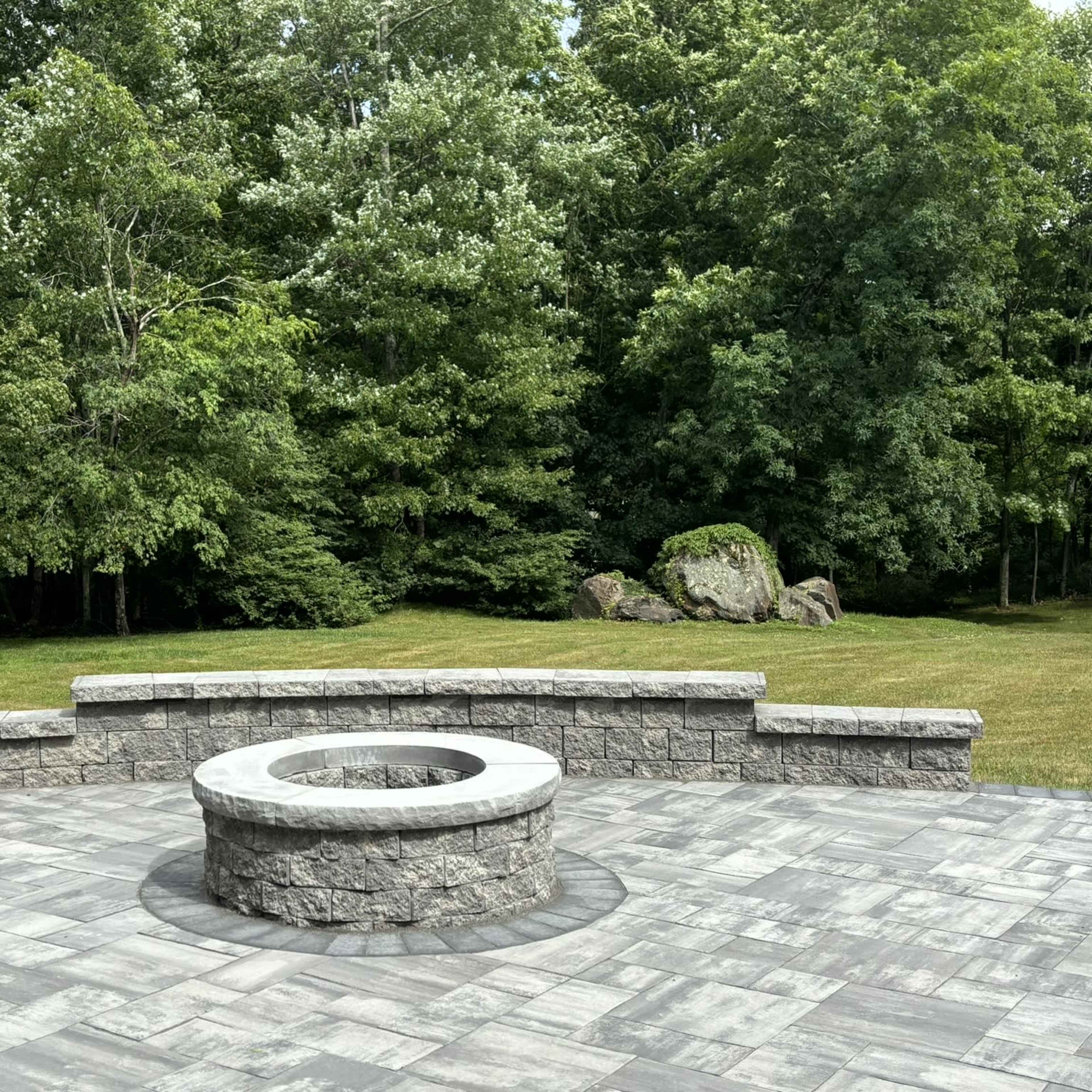 A circular stone fire pit is set in the center of a stone patio surrounded by lush green trees.