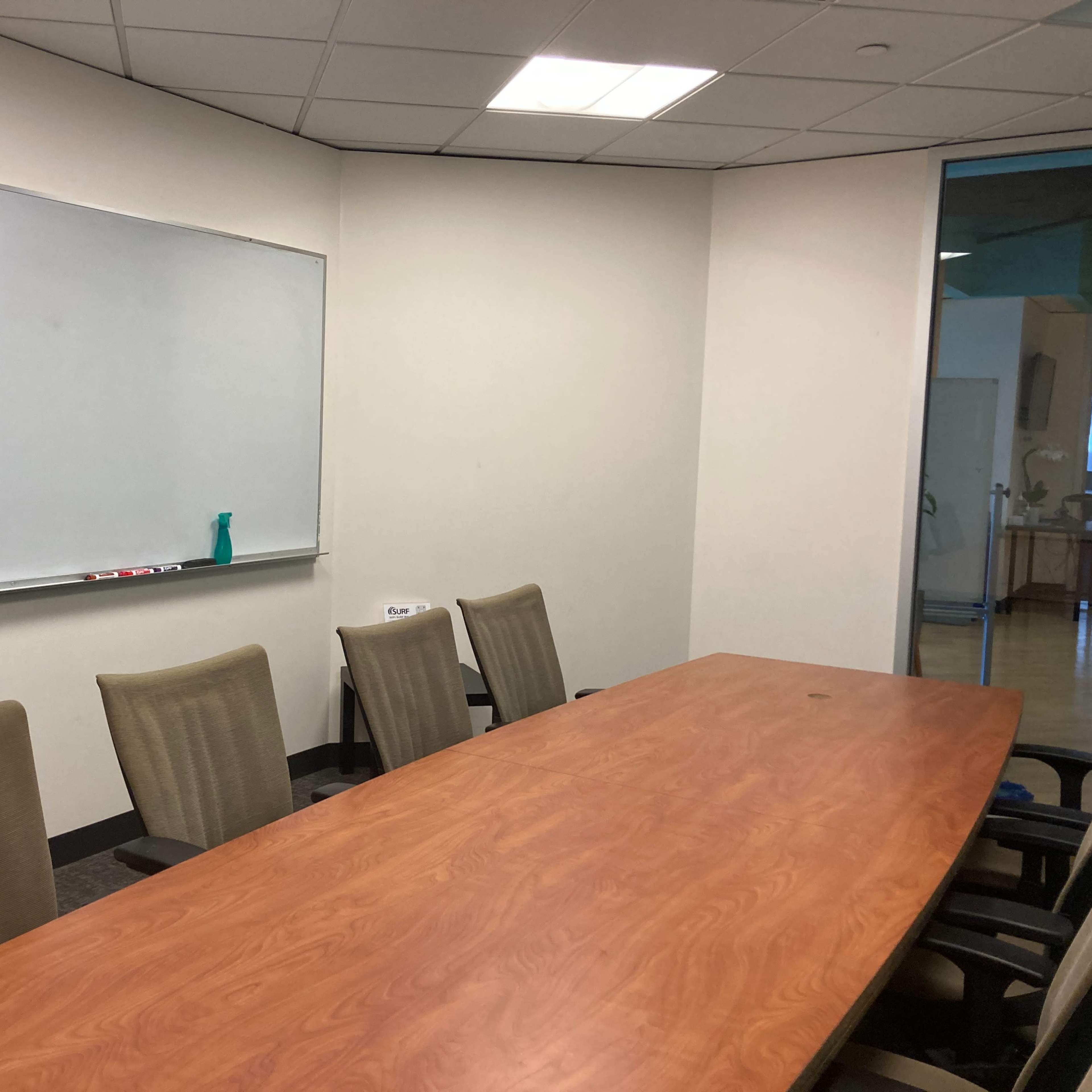 A conference room features a long wooden table surrounded by five chairs and a whiteboard on the wall.