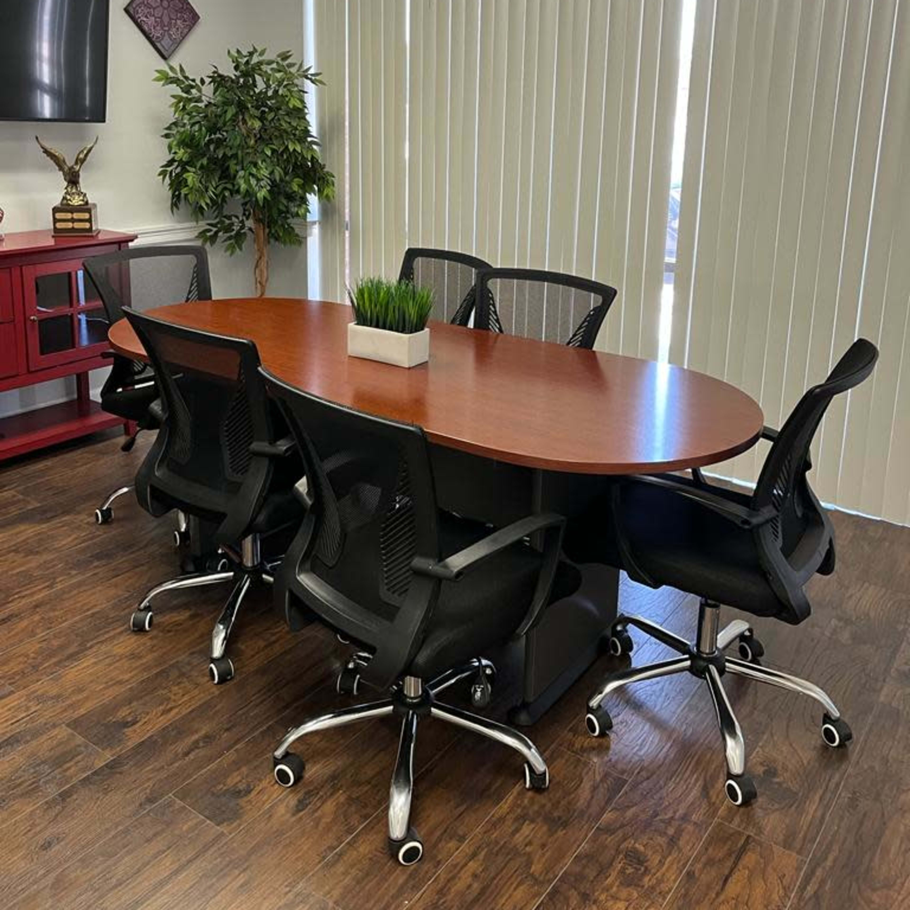 A conference room features a large oval table surrounded by five black office chairs, with a plant centerpiece and a wall-mounted television.