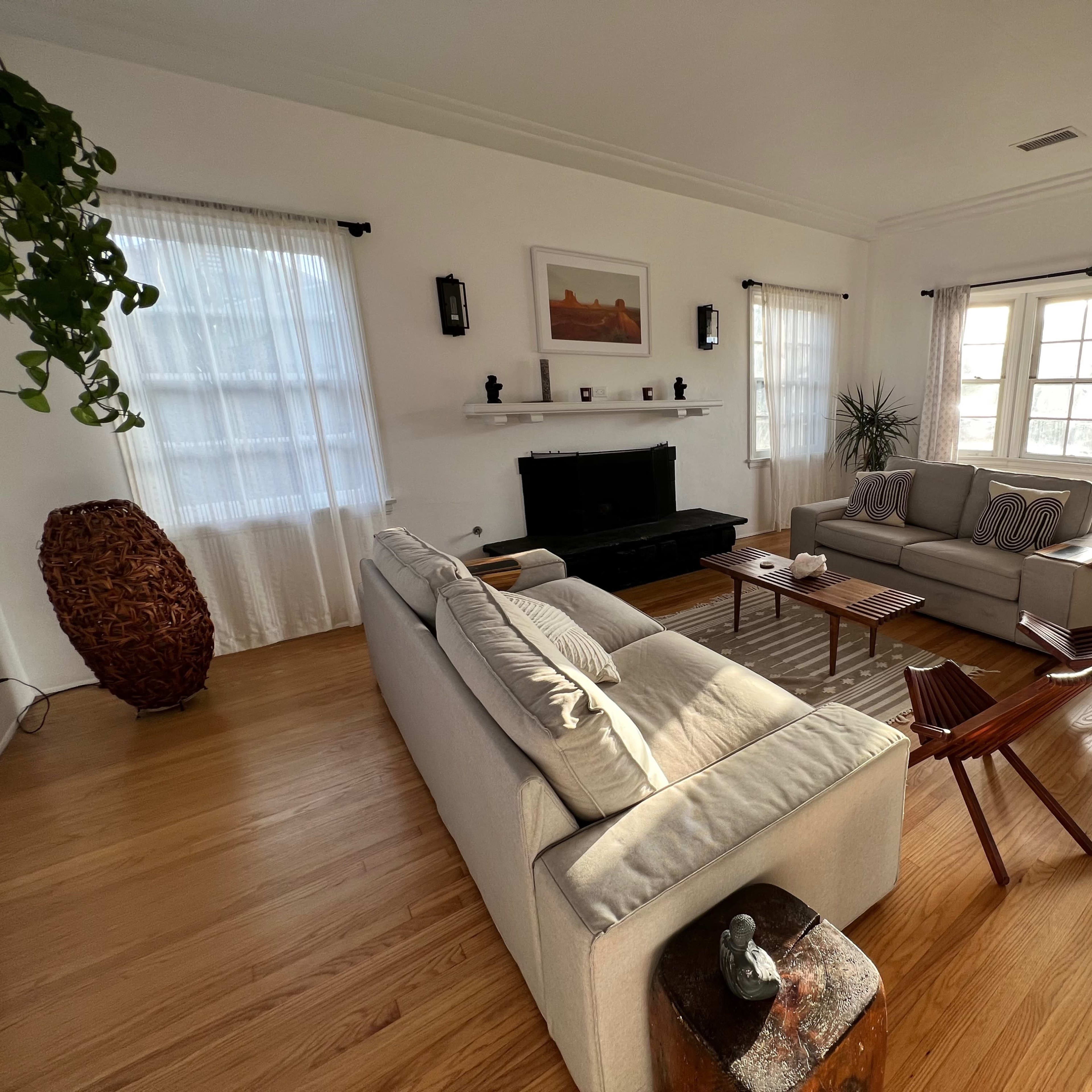 A bright living room features a light-colored sofa, two armchairs, a coffee table, and a fireplace, all set against large windows and wooden floors.