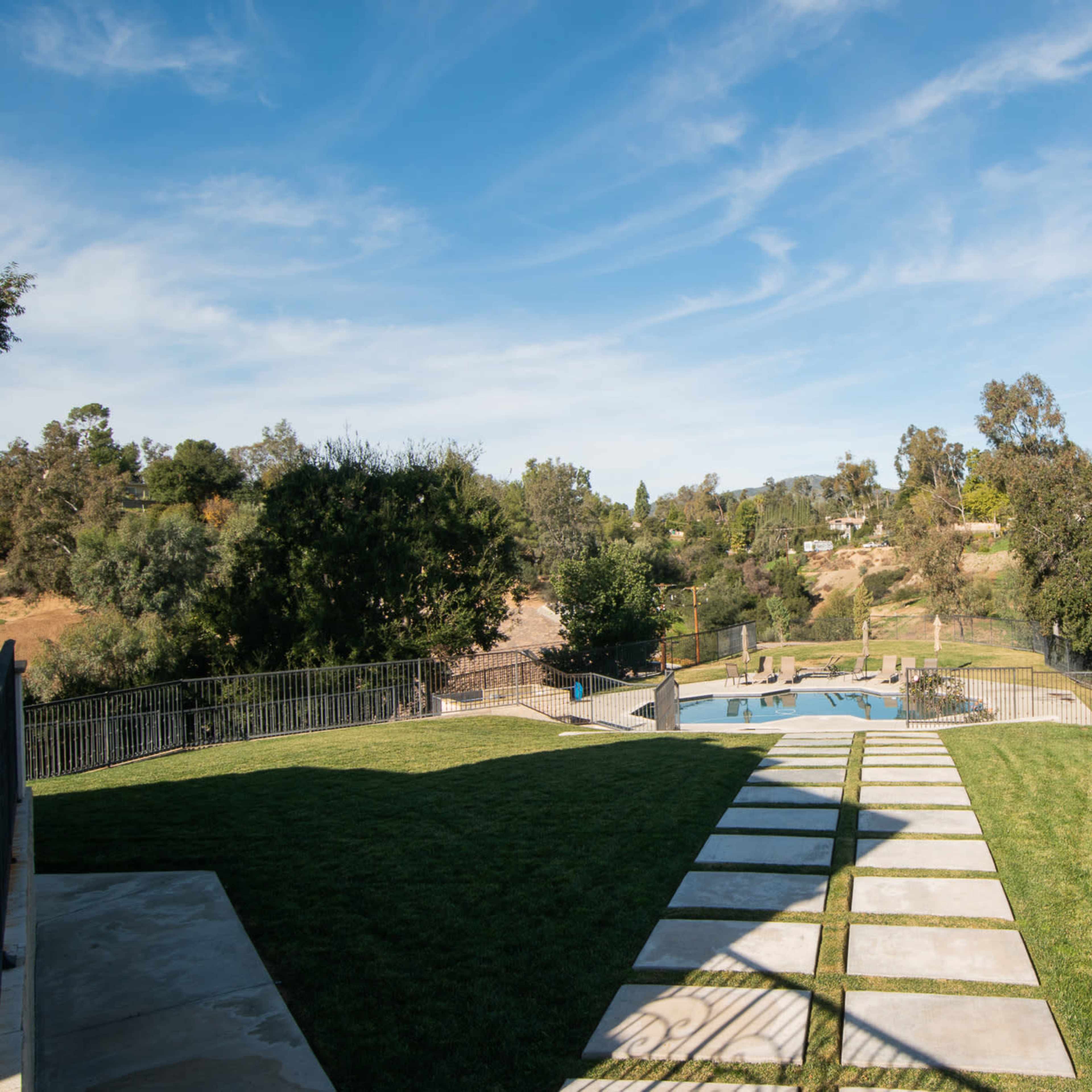 A grassy yard features stone pathways leading to a swimming pool surrounded by trees and hills in the background.