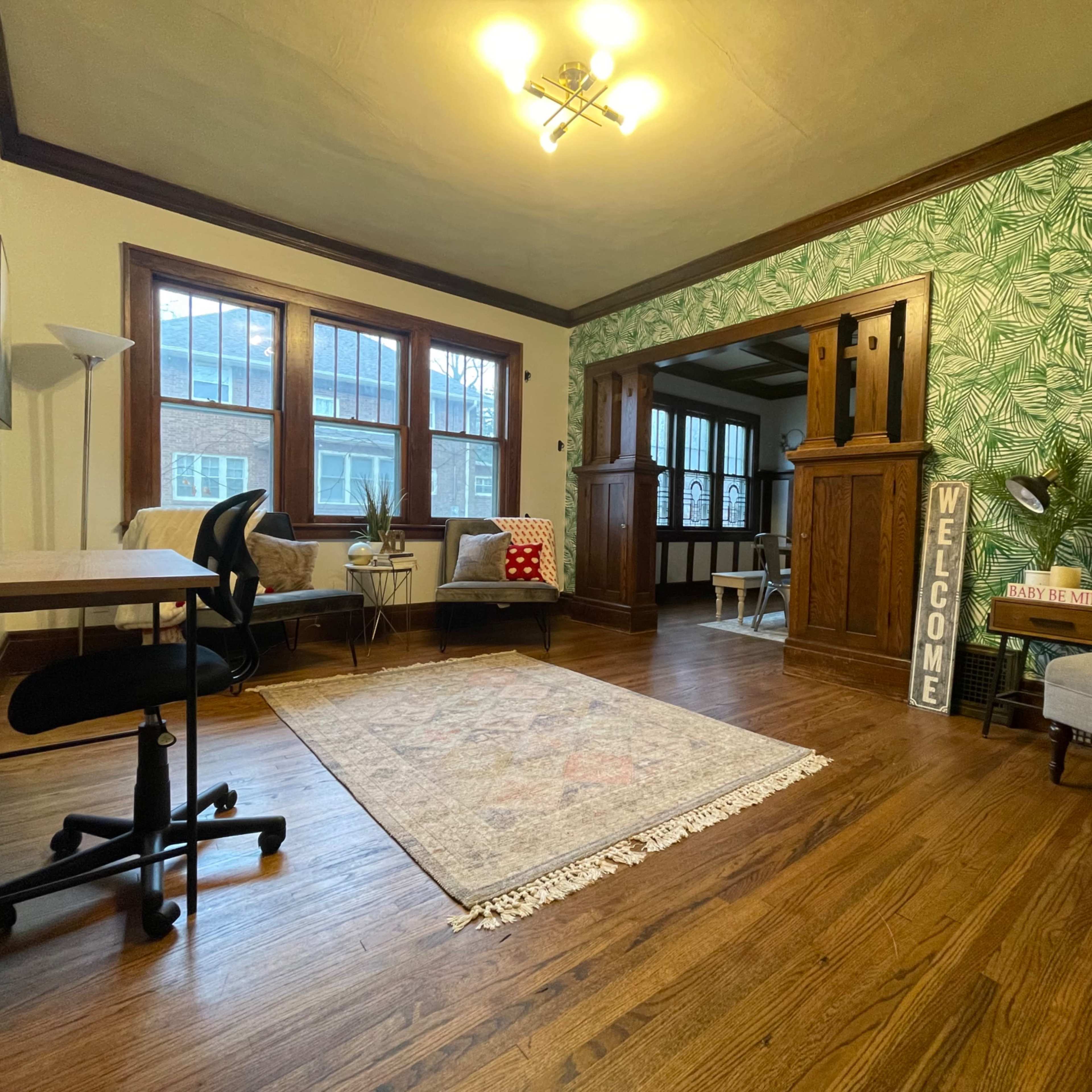 A living room with hardwood floors, a patterned area rug, a desk and chair, and tropical-themed wallpaper.