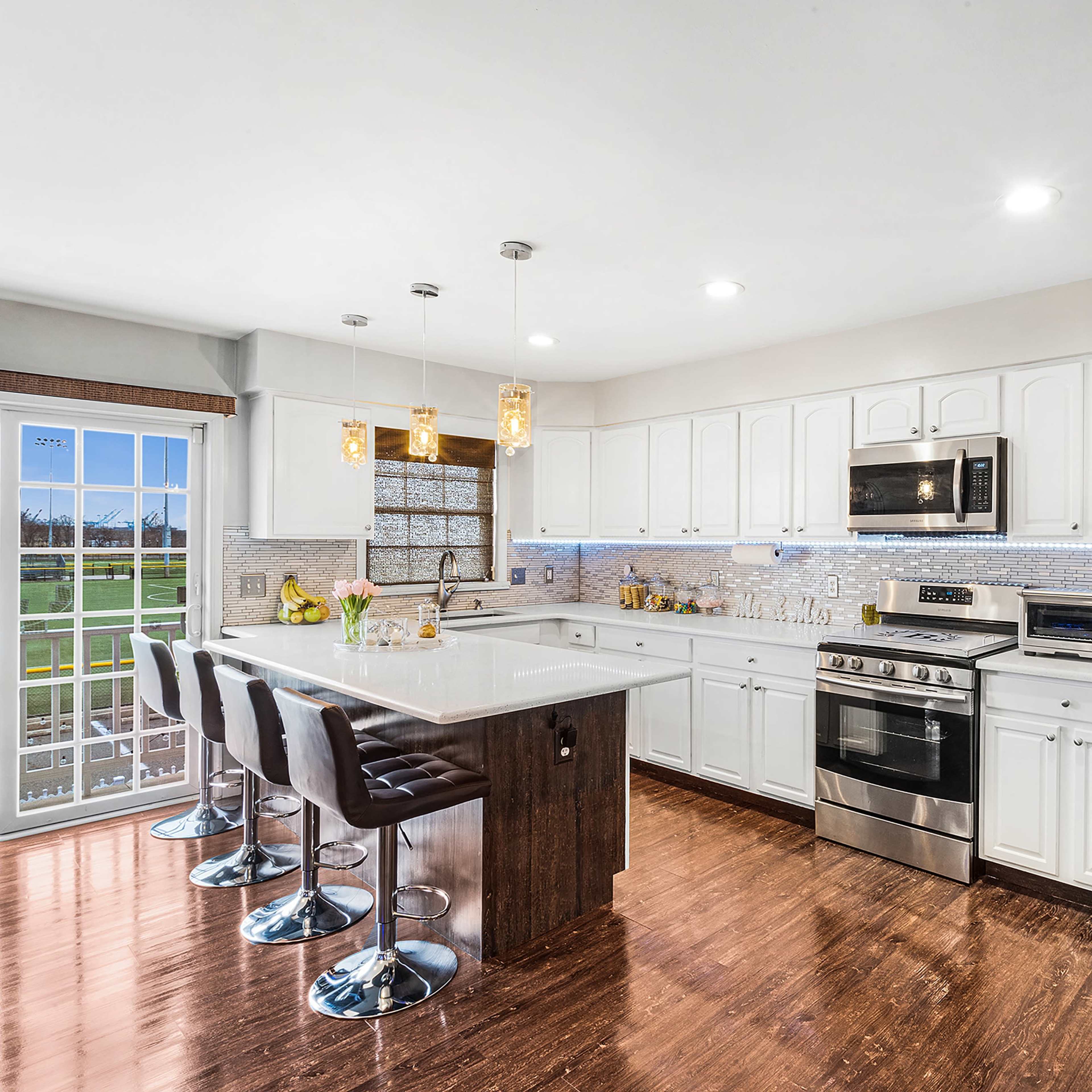 A modern kitchen features white cabinetry, stainless steel appliances, and an island with bar stools, illuminated by pendant lights.