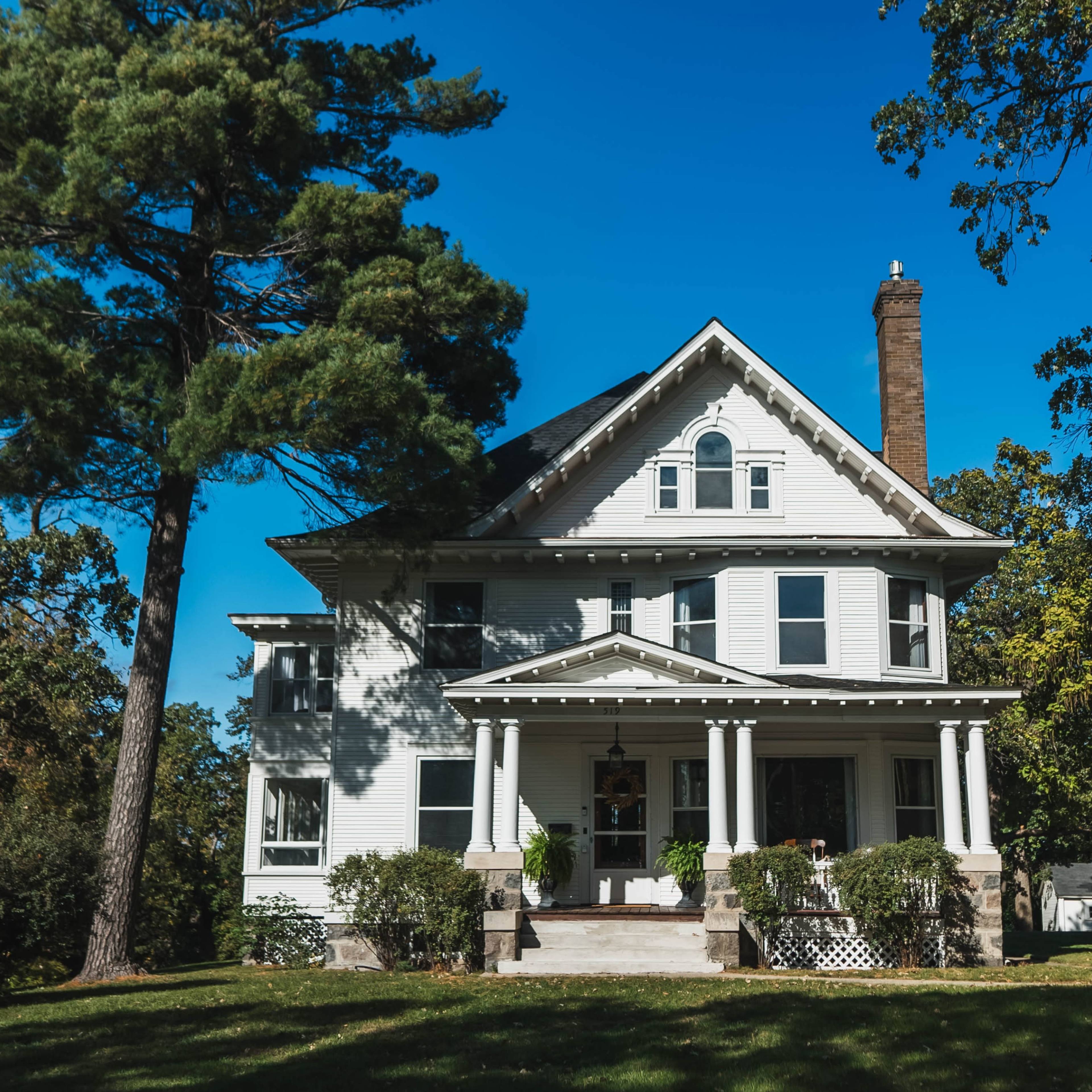 A large, two-story white house with a porch and tall trees stands on a grassy lawn under a clear blue sky.