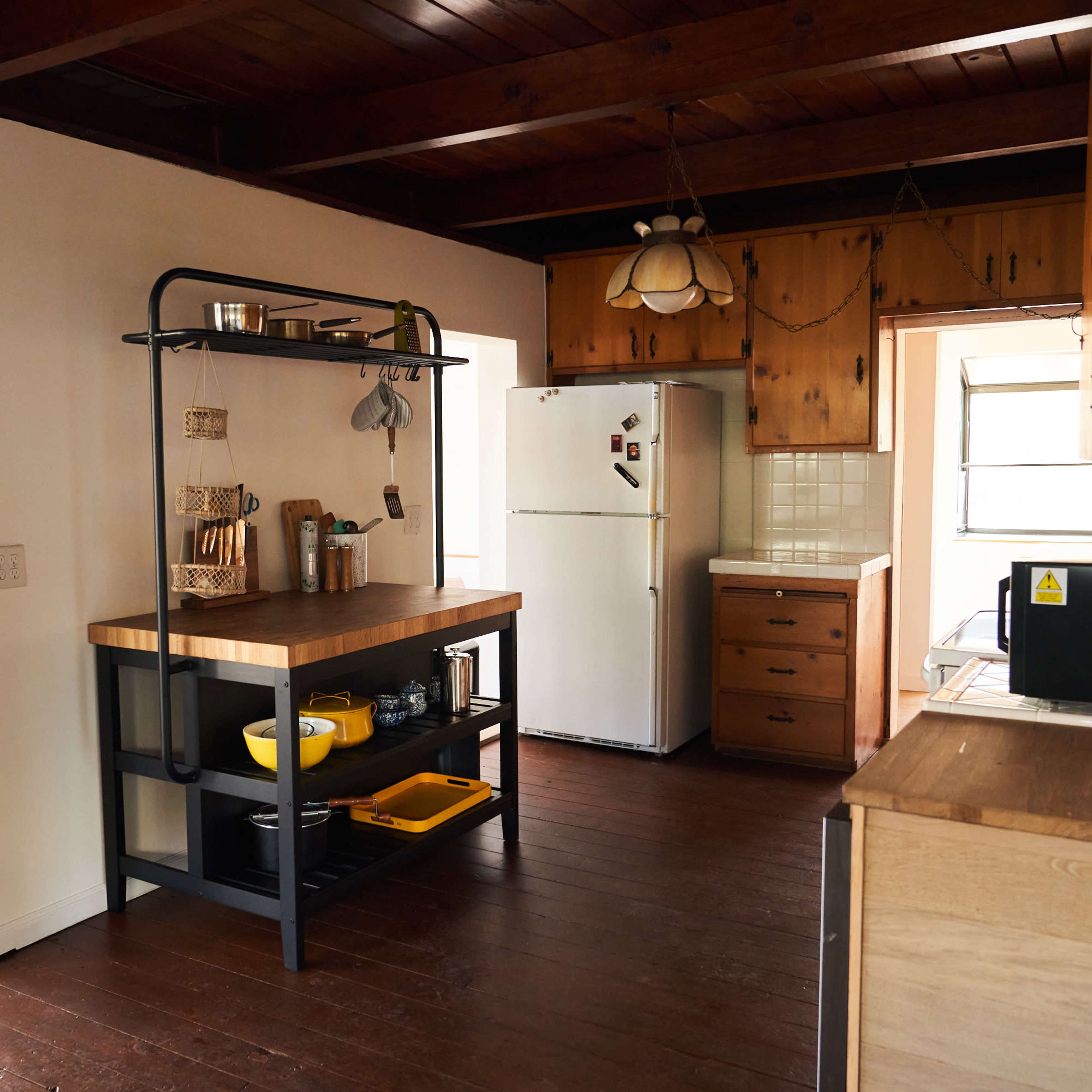 A kitchen features wooden floors, a central island with a black frame, and various appliances, including a refrigerator and microwave, against wooden cabinetry.