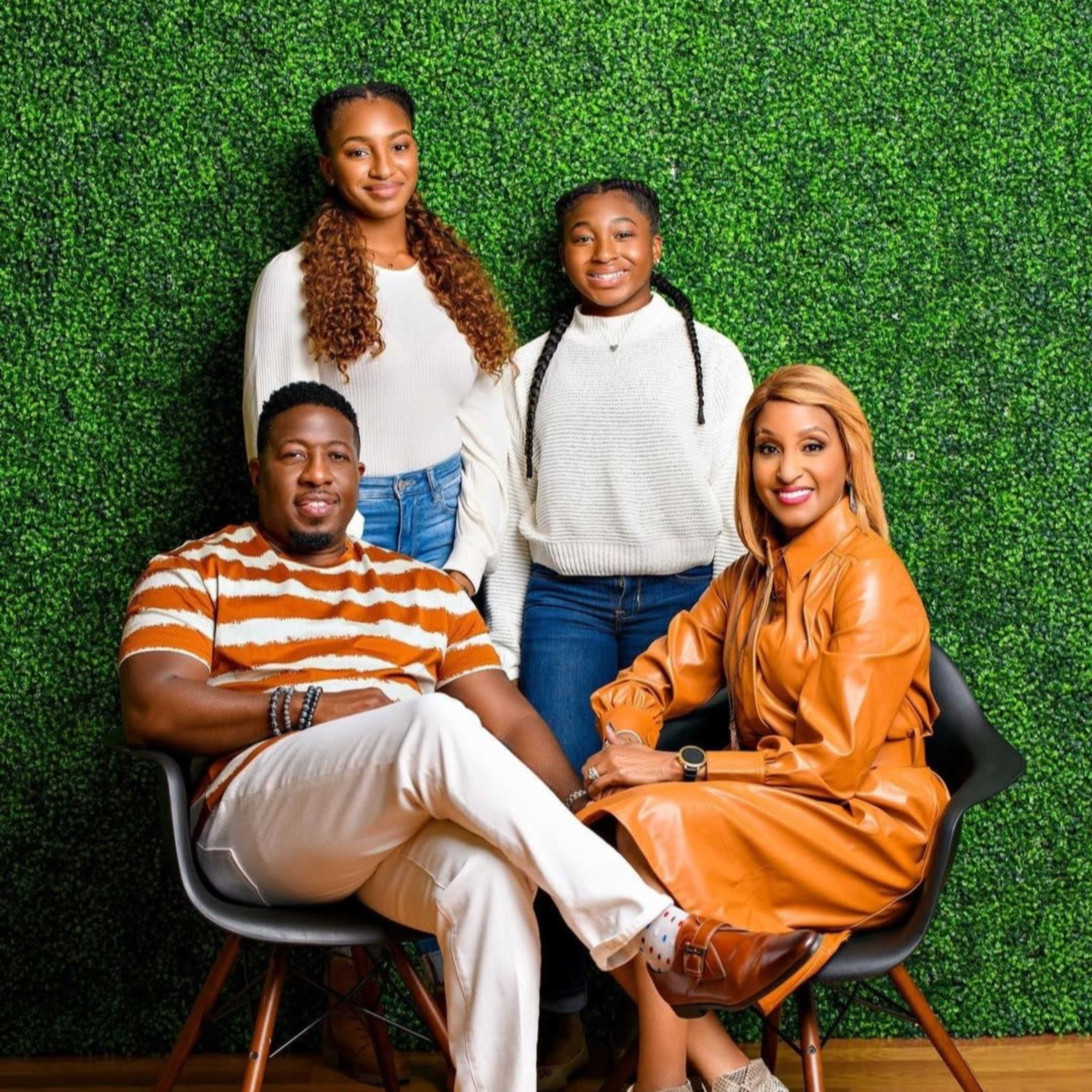 Four people pose for a photo in front of a green artificial hedge wall, with one man seated in a chair and three women standing behind him.