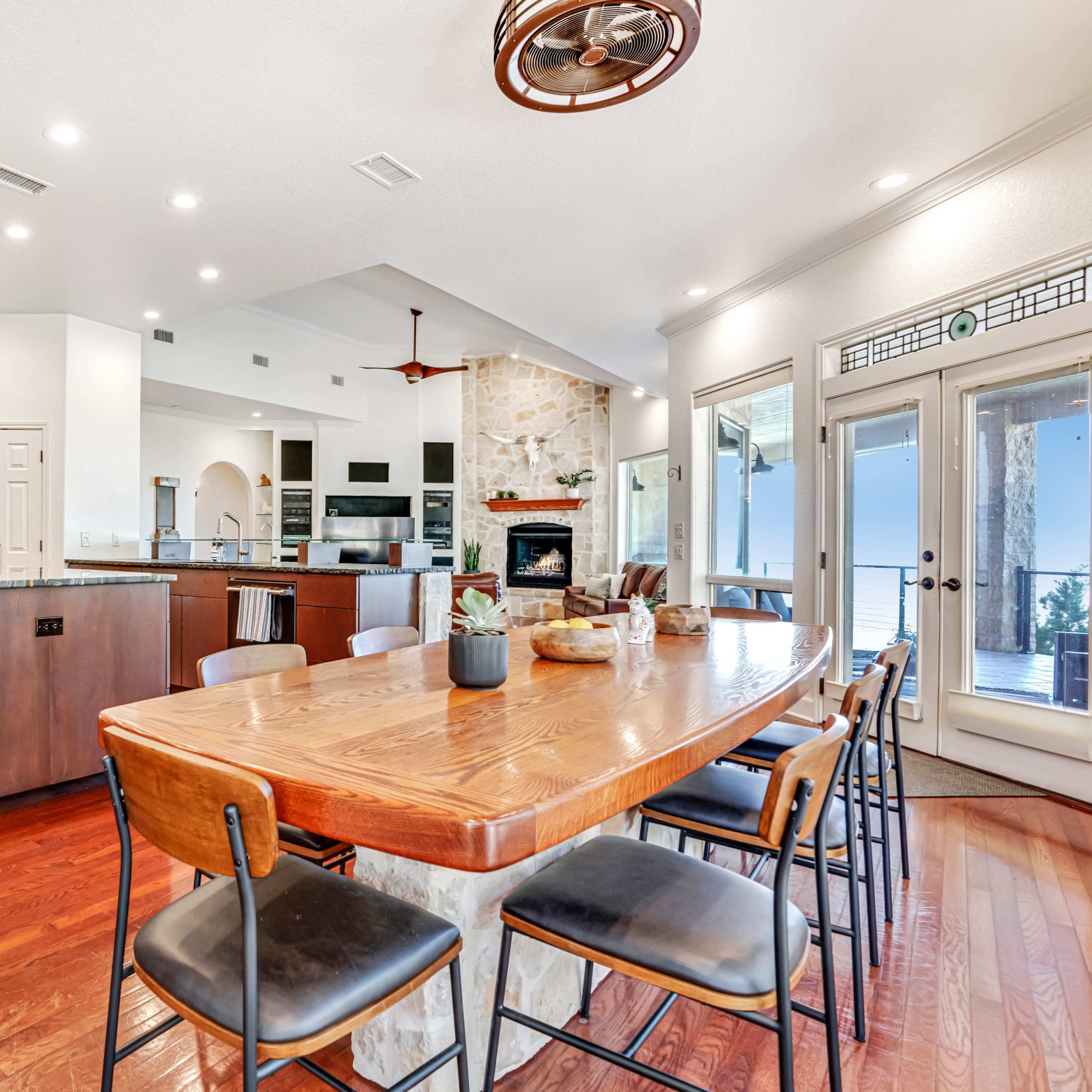 A modern kitchen features a large island with dark cabinetry, a wood dining table, and an open layout leading to a living area with a stone fireplace.