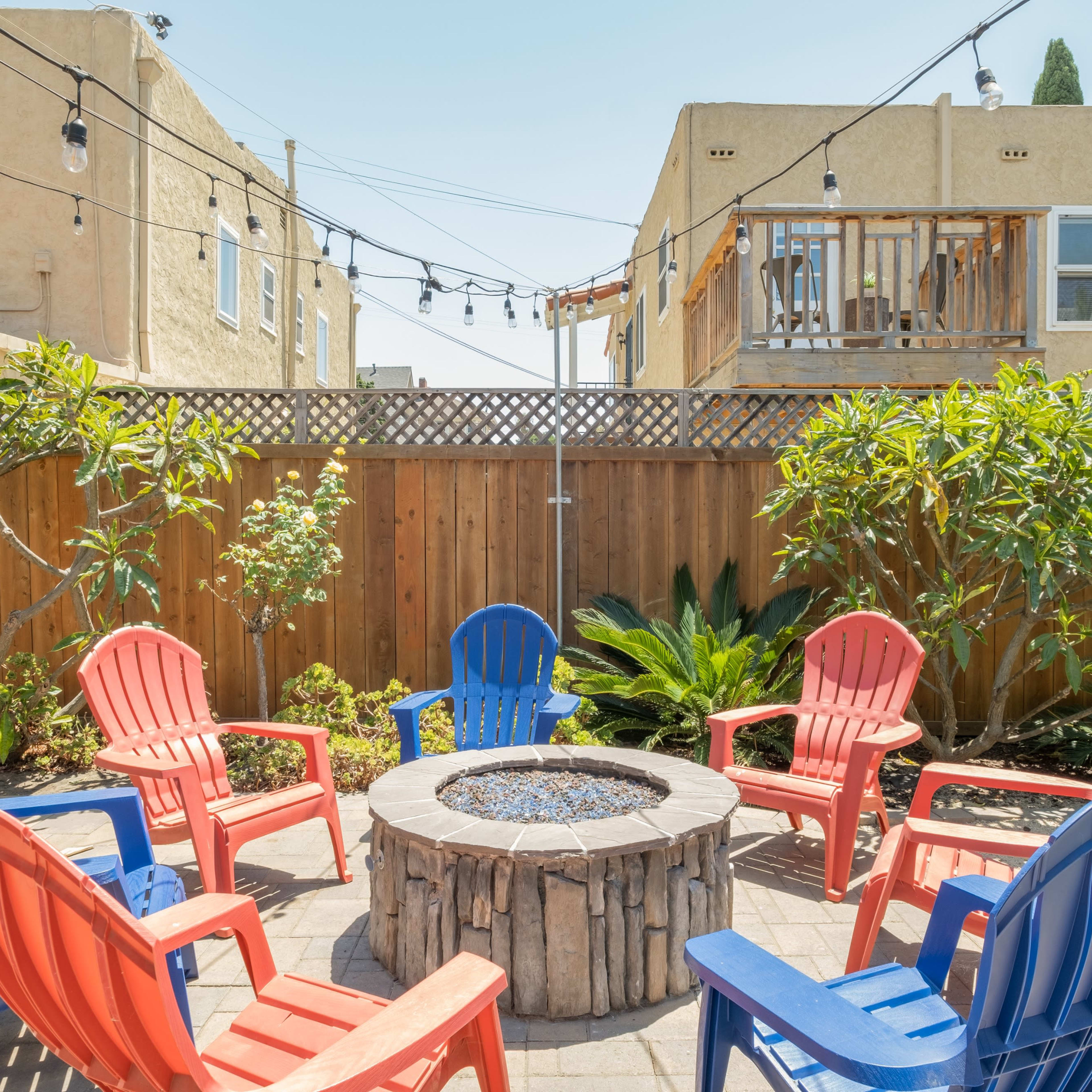 A circular arrangement of red and blue chairs surrounds a stone fire pit in a landscaped backyard with string lights overhead.