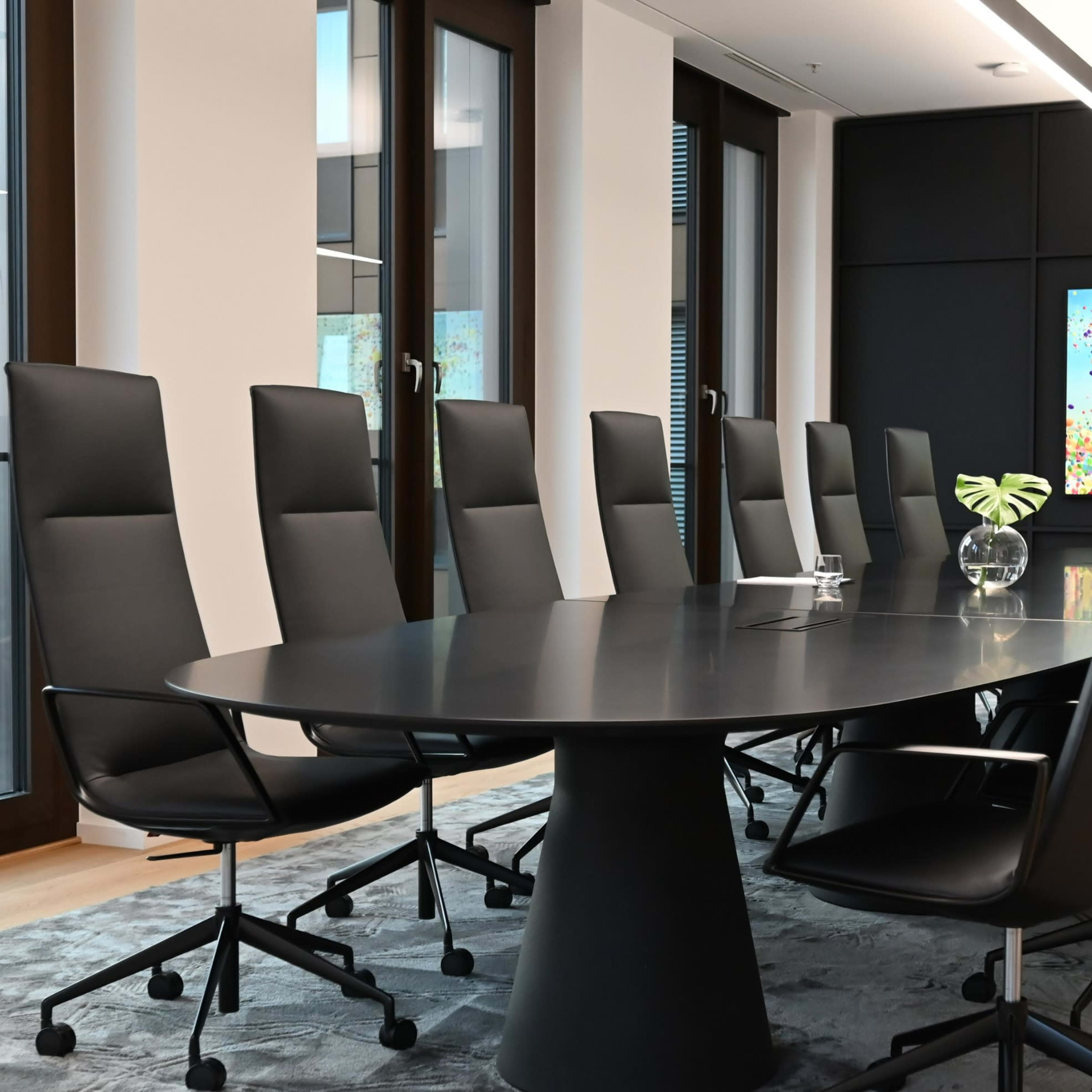 A modern conference room features a large oval table surrounded by ergonomic chairs, with floor-to-ceiling windows and contemporary artwork on the walls.