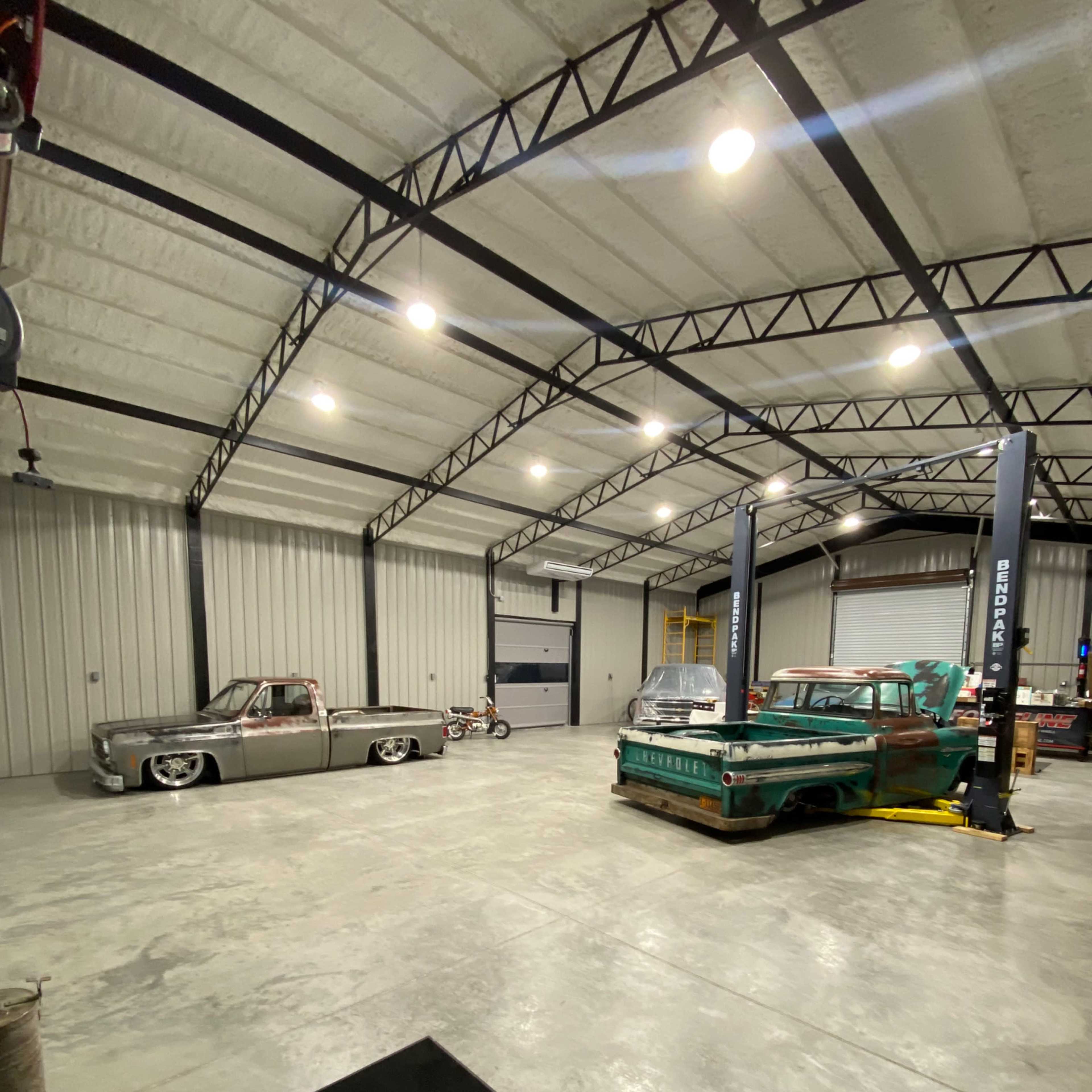 An industrial garage features several classic cars, including a silver pickup and a green vehicle, with a vehicle lift in the center and concrete flooring.