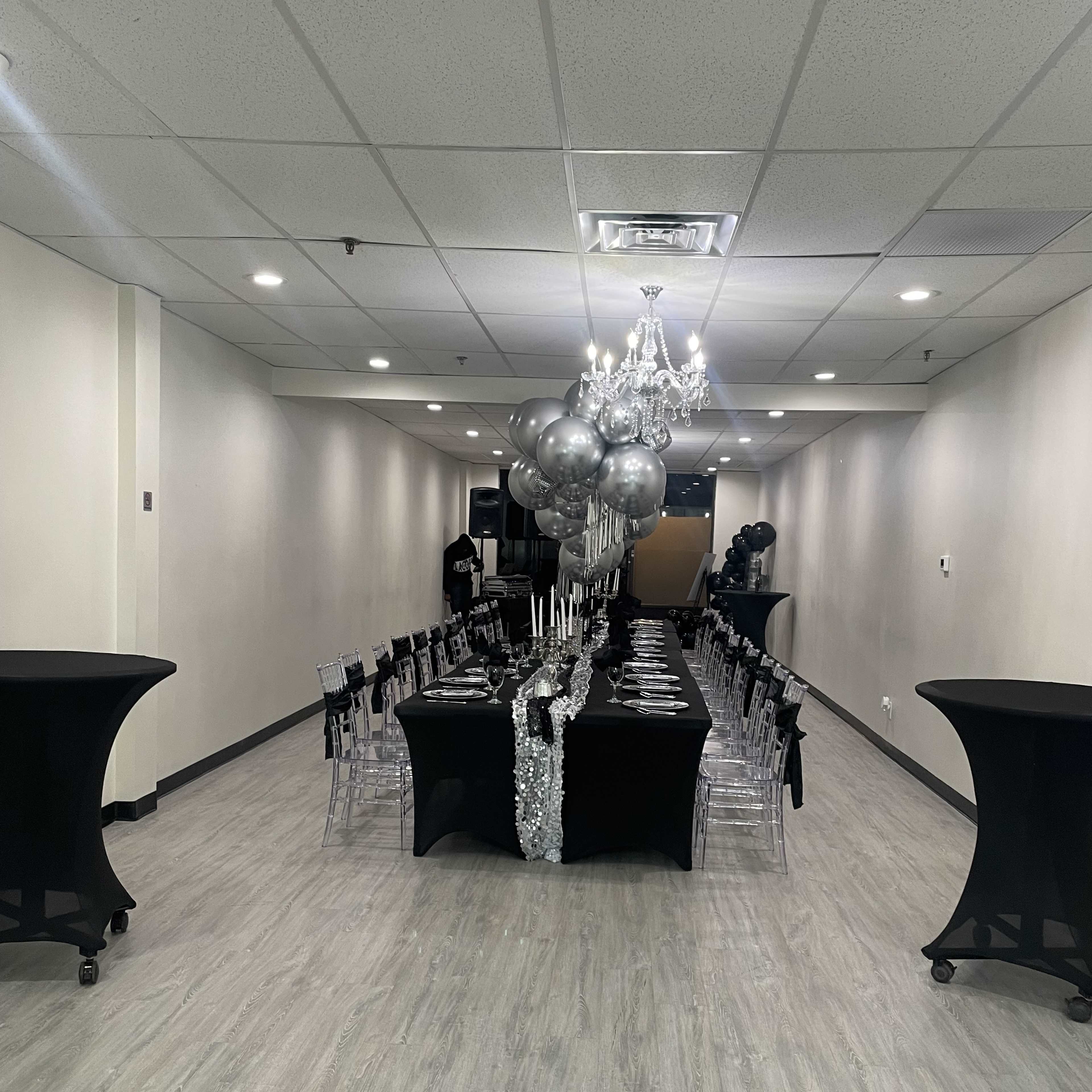 A long dining table is set with black tablecloths and silver decorations in a spacious, well-lit event room.