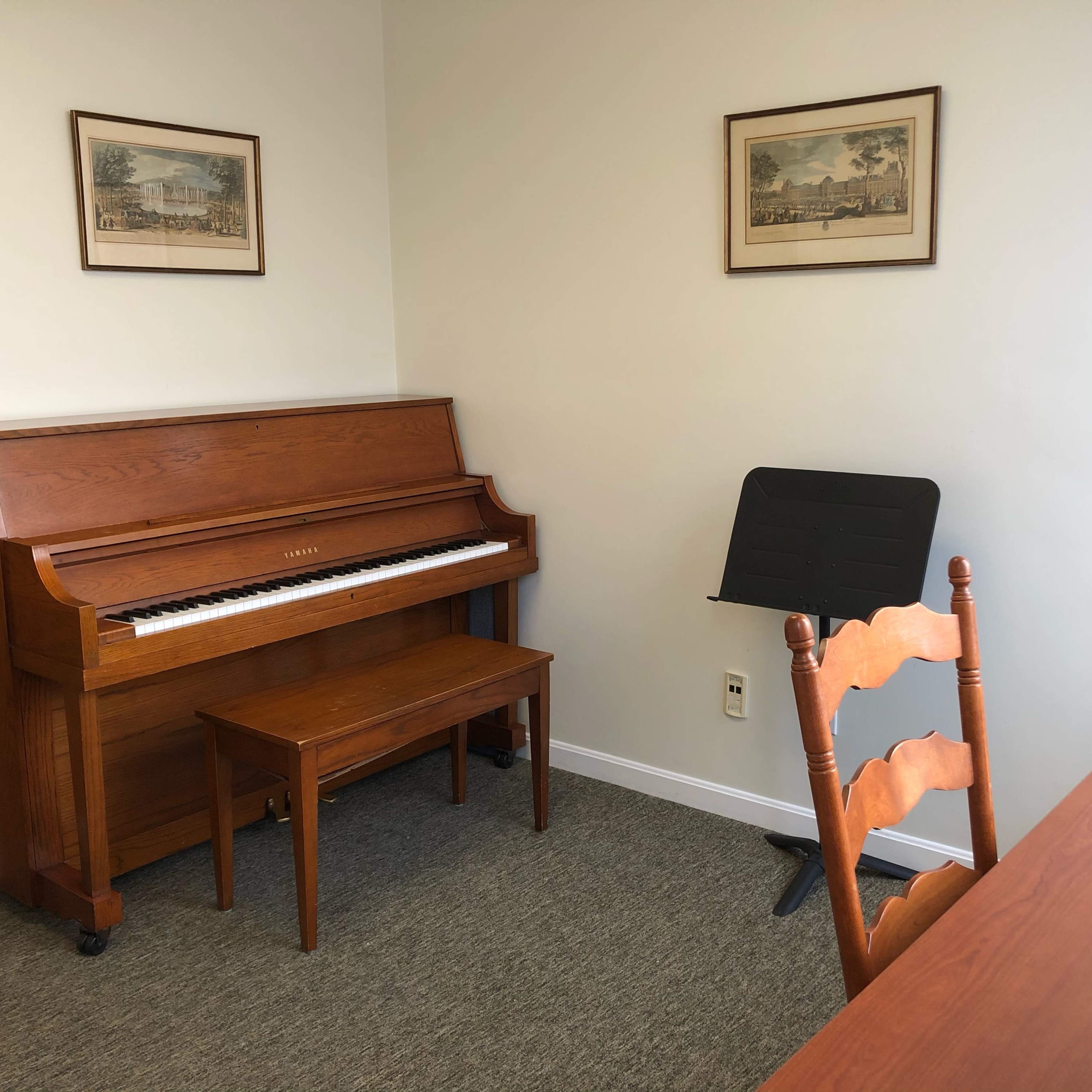 A wooden piano is positioned against a wall in a small, well-lit room featuring a chair and a music stand.