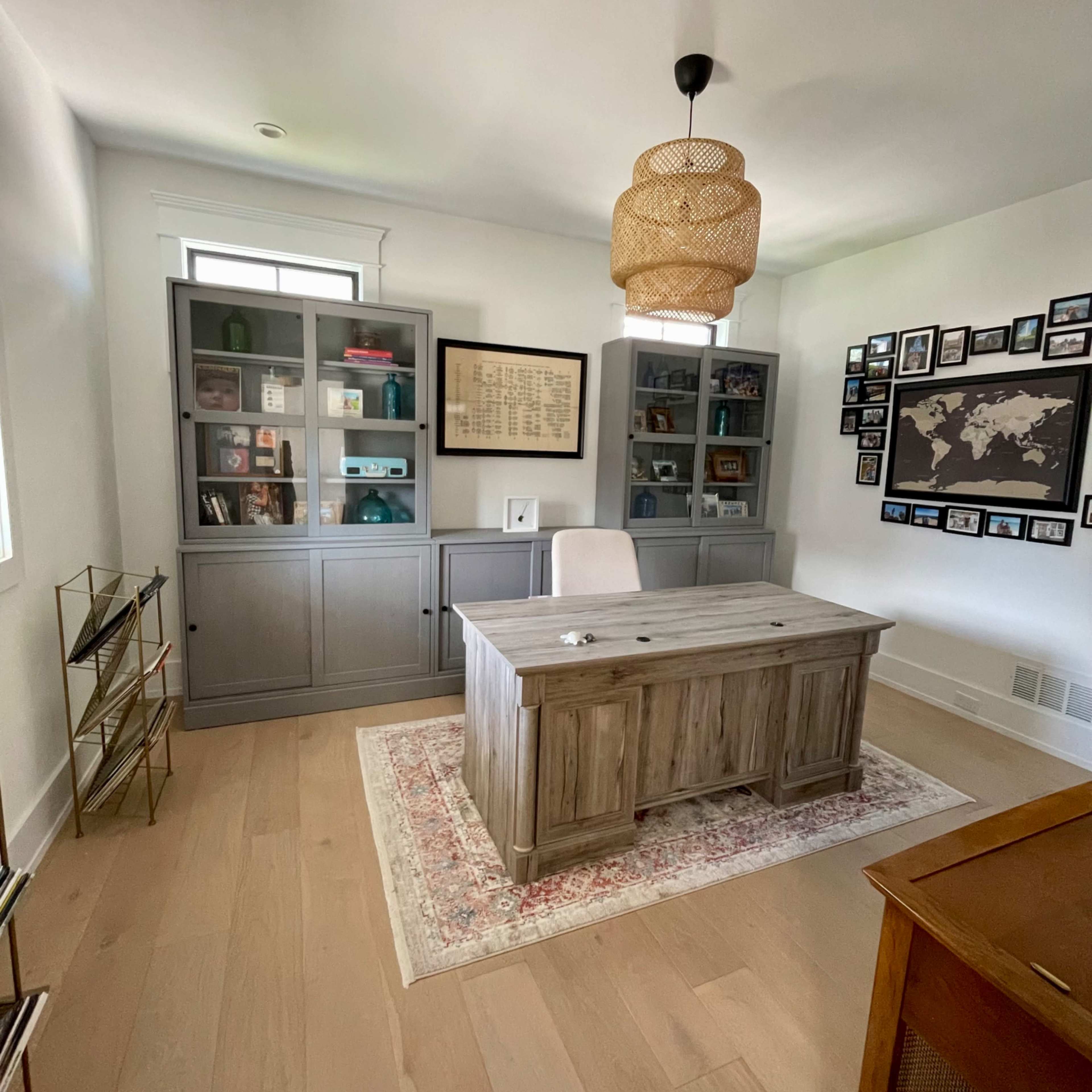 A modern office with a wooden desk, a light fixture, built-in shelves containing various items, and a wall adorned with framed photographs and a world map.