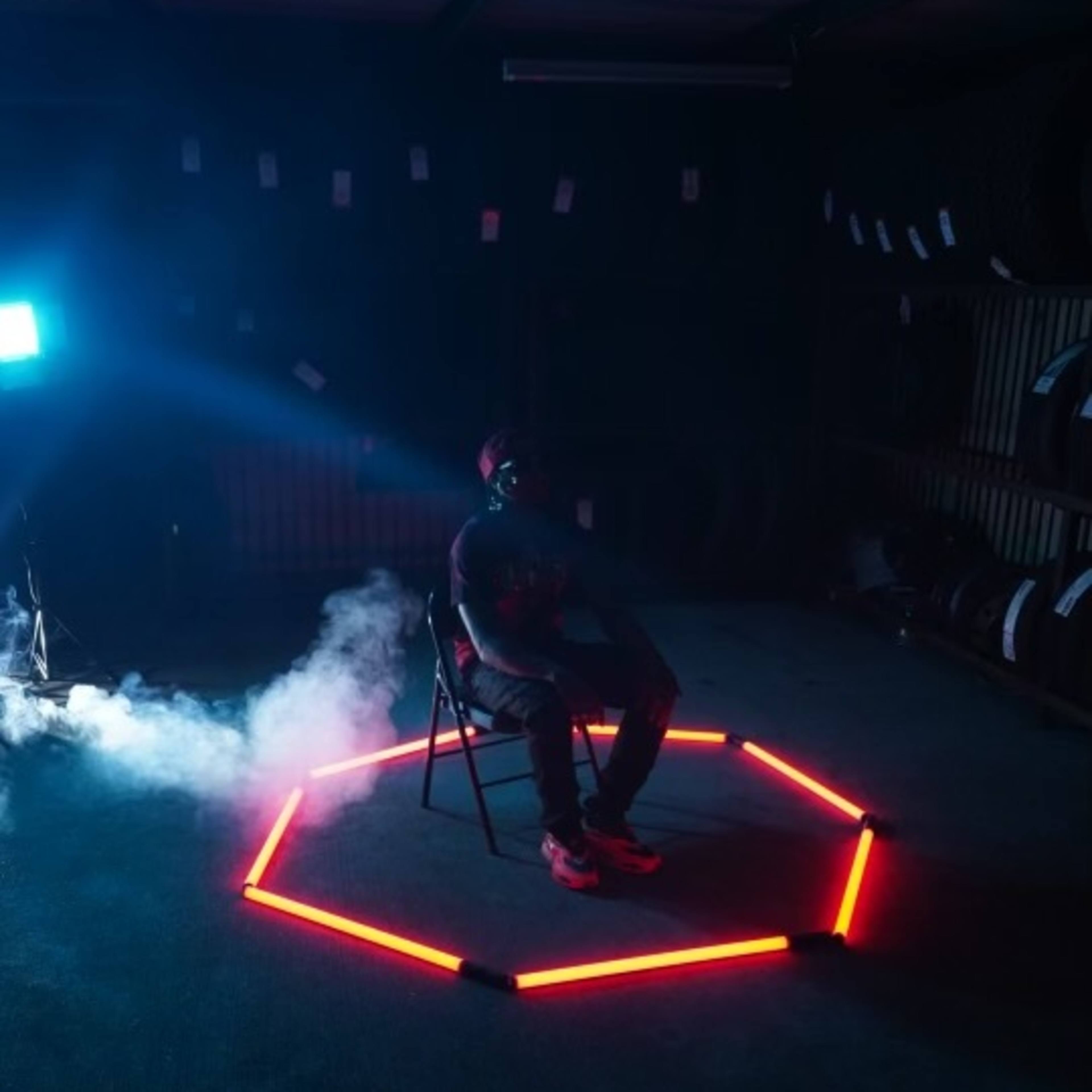 A person sits on a chair in the center of an illuminated hexagonal shape made of red lights, surrounded by smoke in a dimly-lit room.