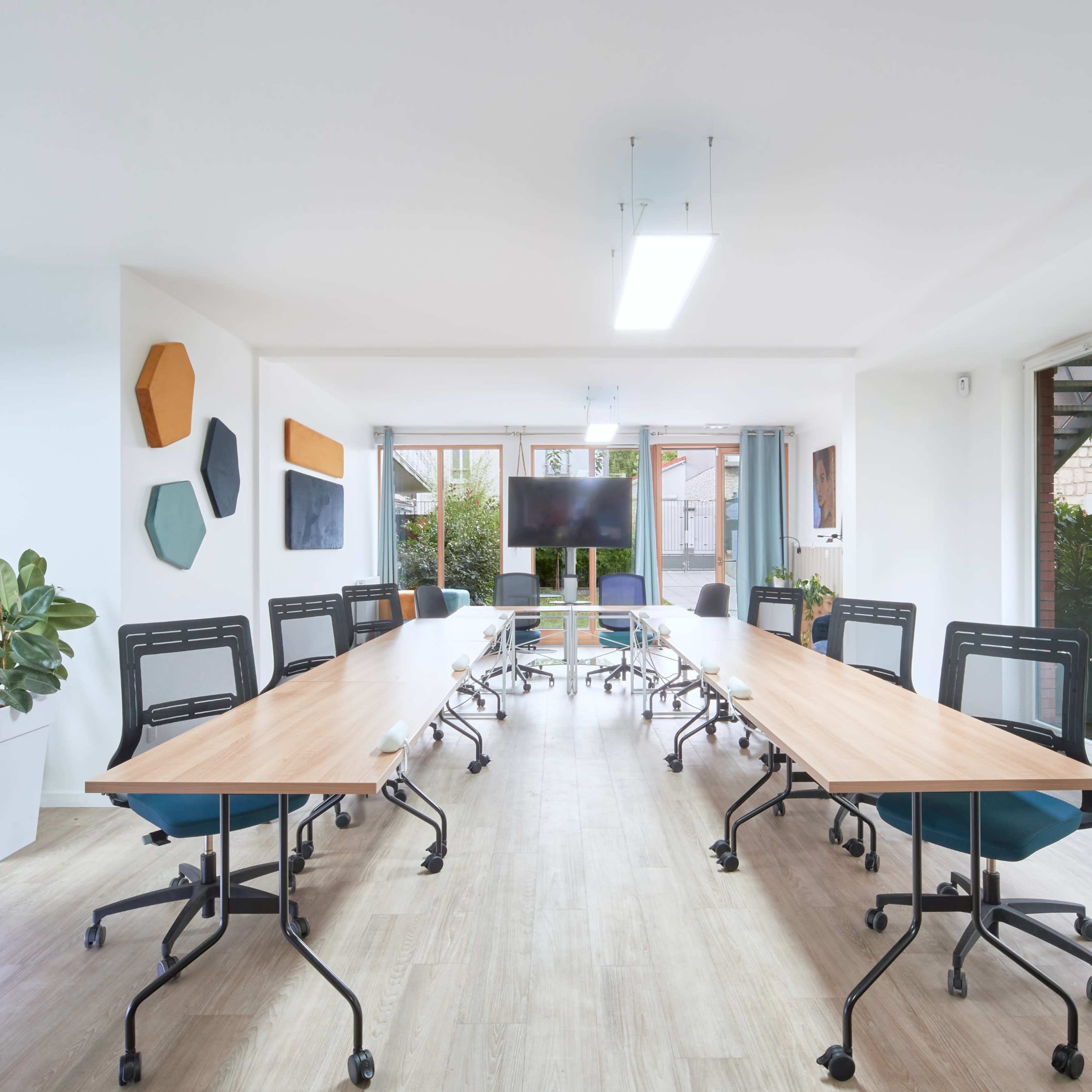 A modern conference room features a long table surrounded by ergonomic chairs, with large windows providing natural light and greenery visible outside.