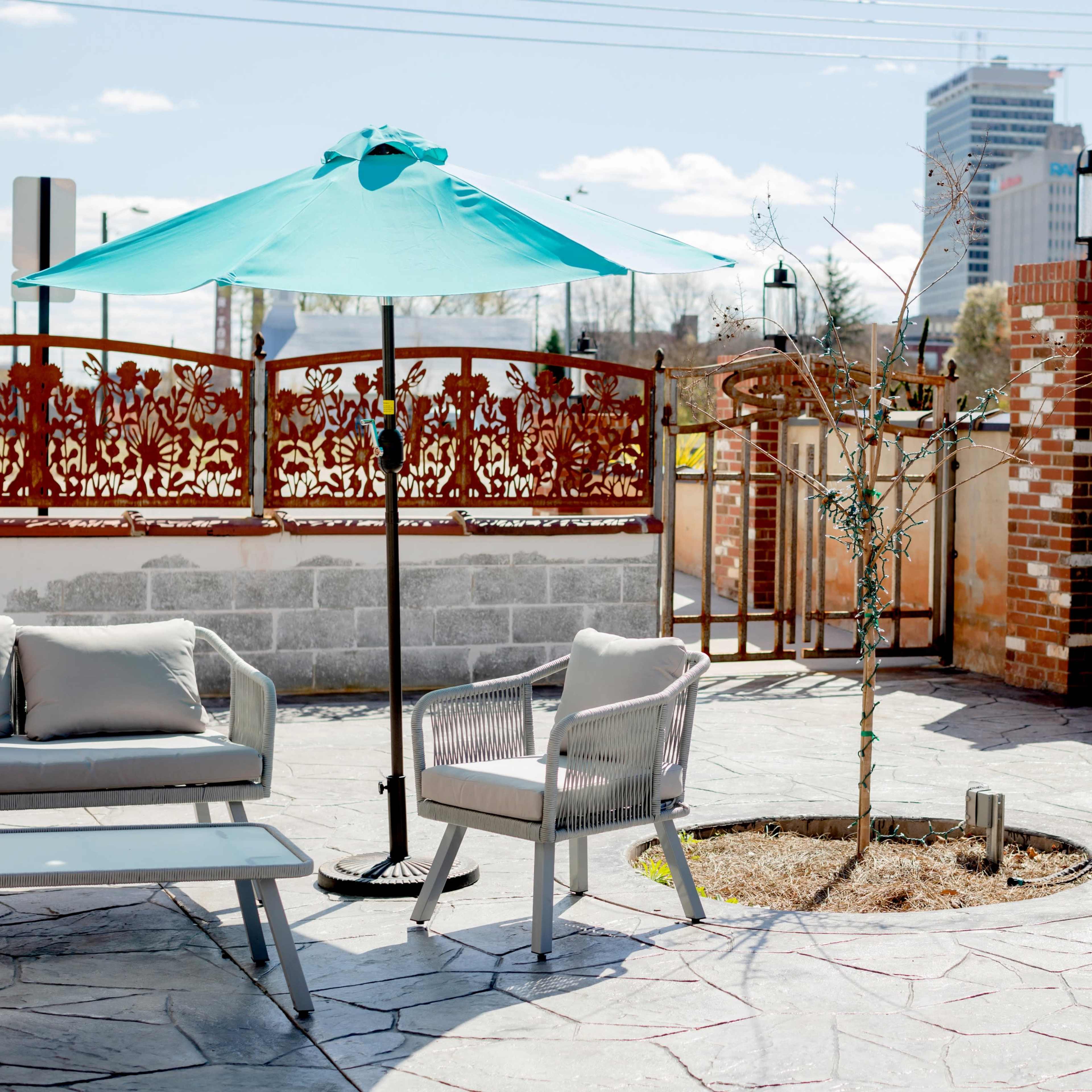 A rooftop patio with a teal umbrella, a light-colored sofa set, and a small tree planted in a circular garden area, surrounded by a decorative wall and a city skyline in the background.