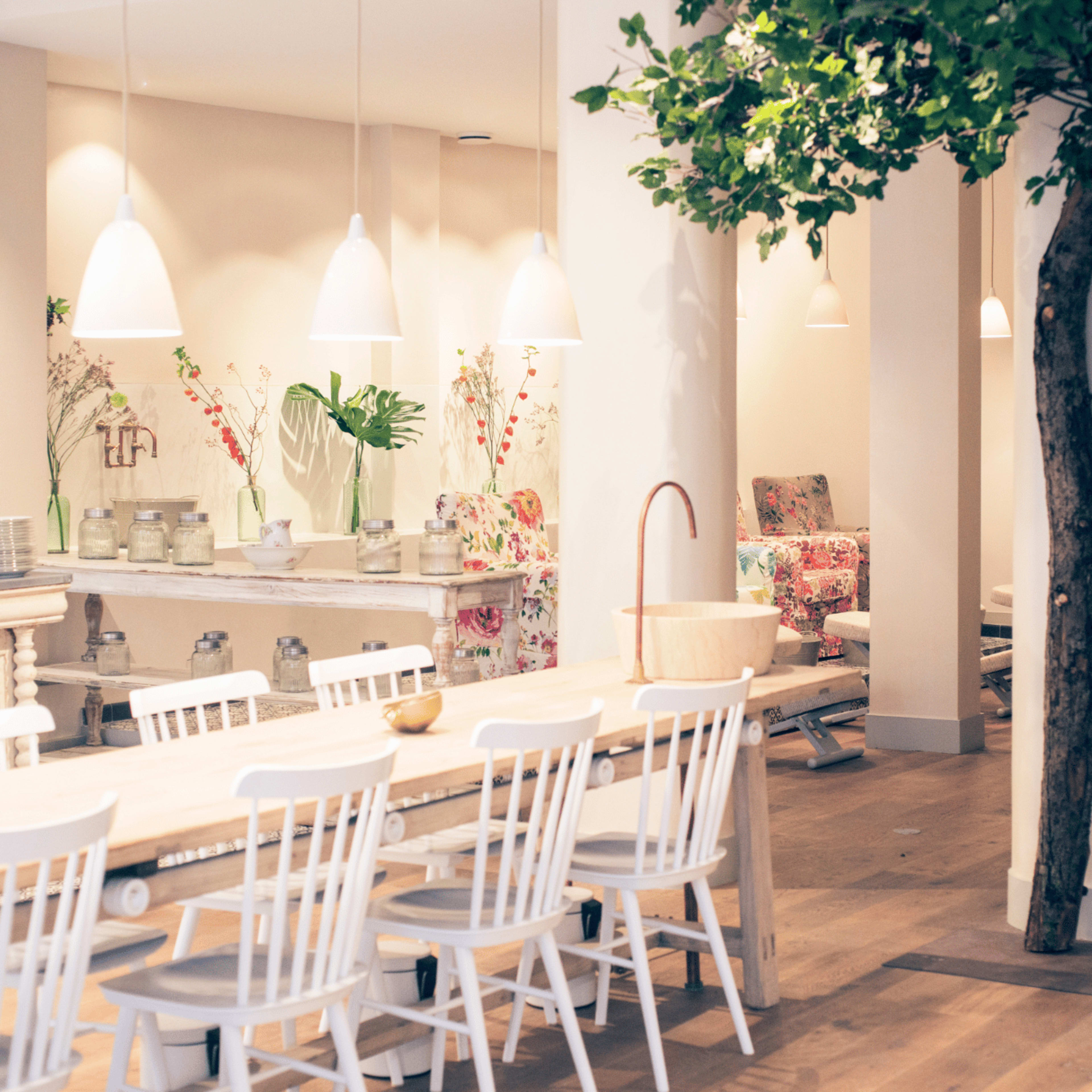 A spacious dining area features a long wooden table surrounded by white chairs, with a tree and decorative elements in a softly lit environment.