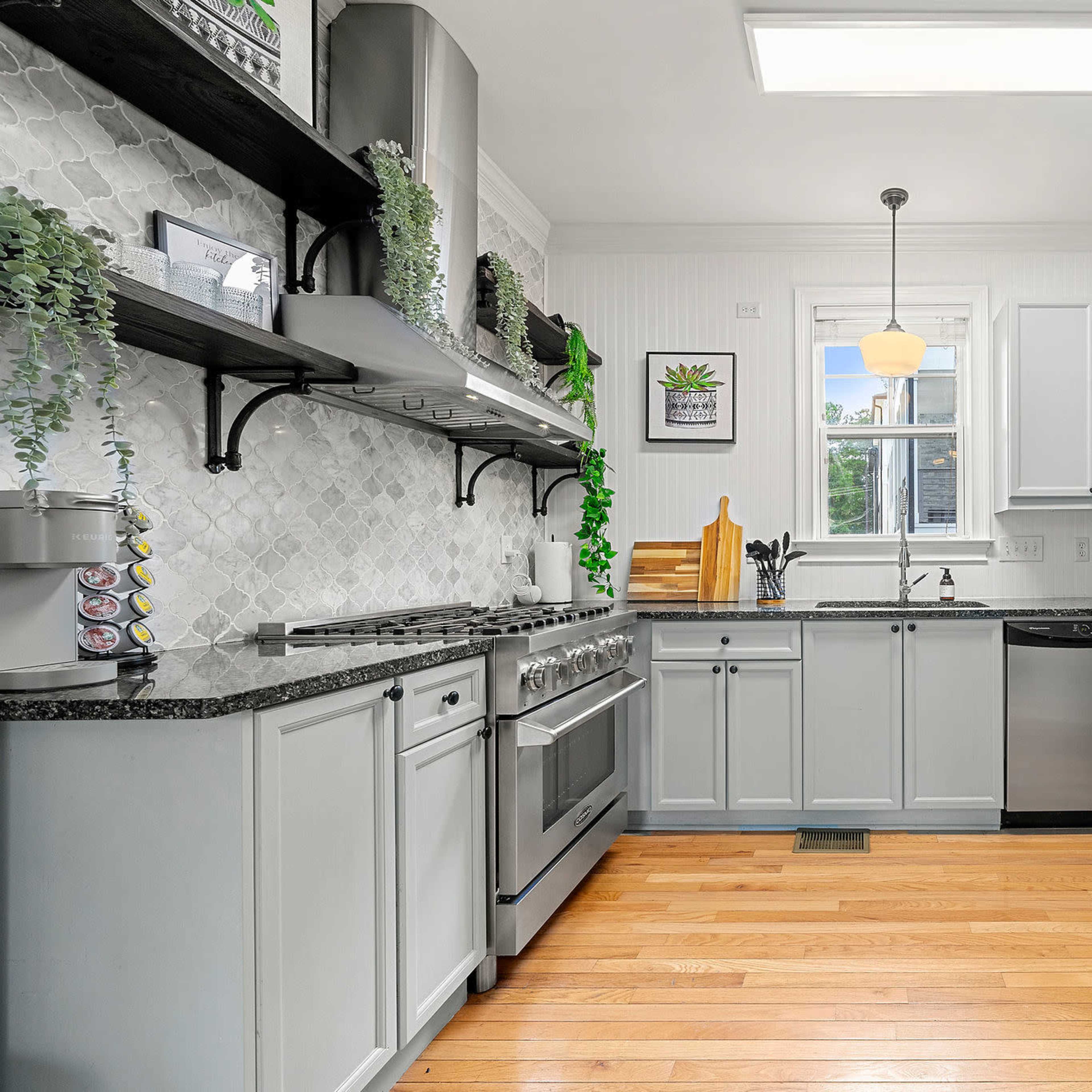 A modern kitchen with gray cabinets, stainless steel appliances, and wooden flooring, featuring a window and decorative plants on the shelves.