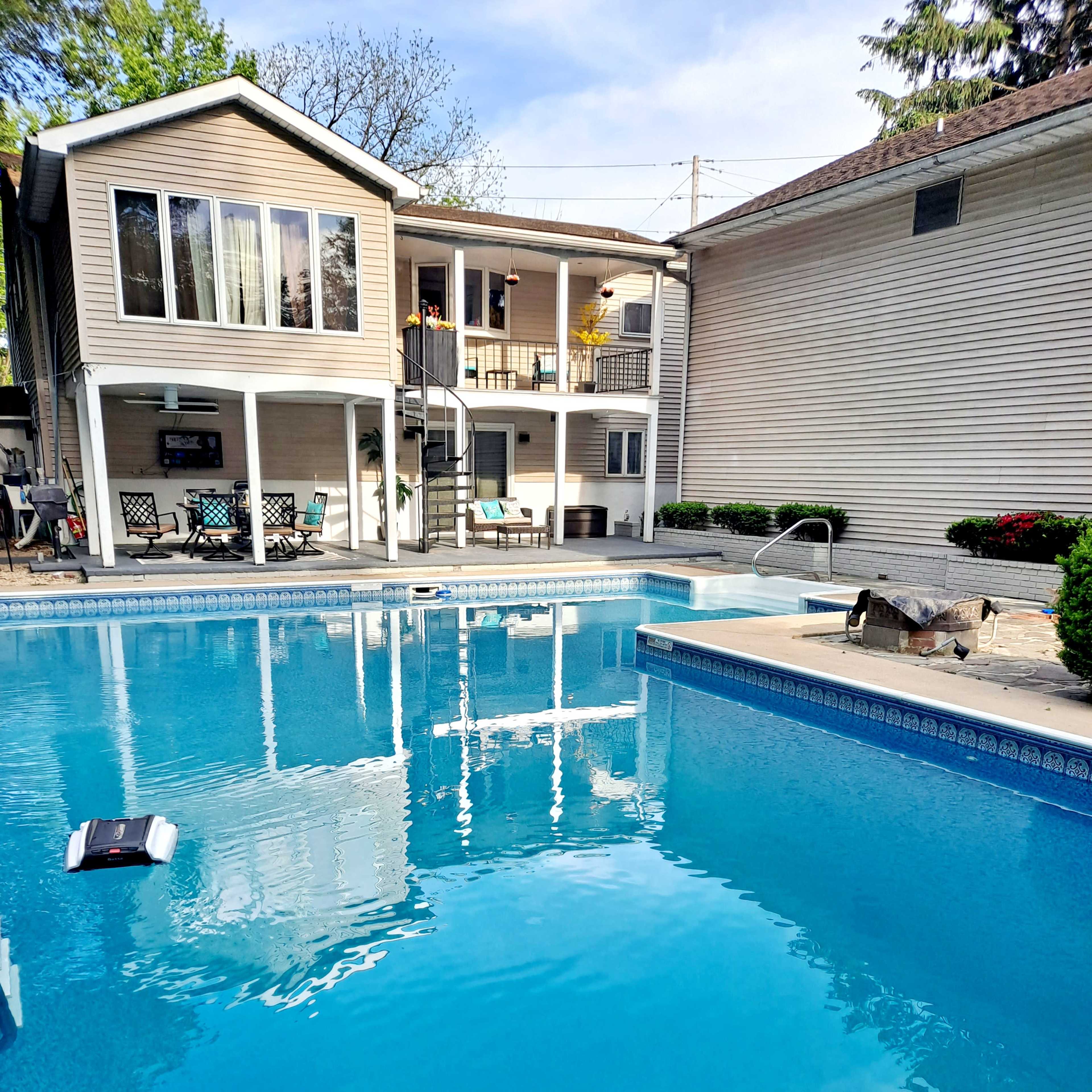 A swimming pool in a backyard with a two-story house and a patio area visible in the background.