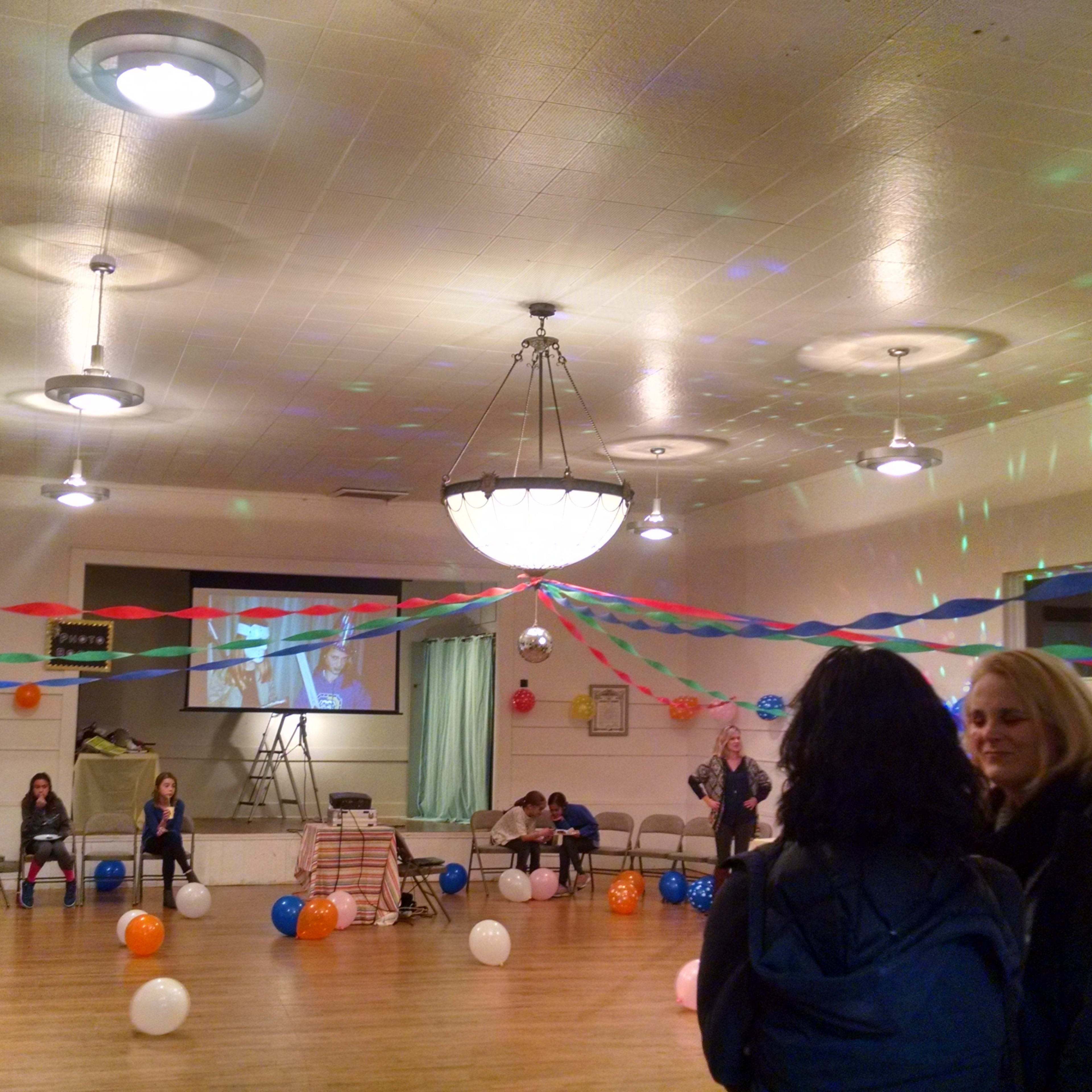 A decorated indoor event space features colorful balloons, streamers, and a few people mingling around.