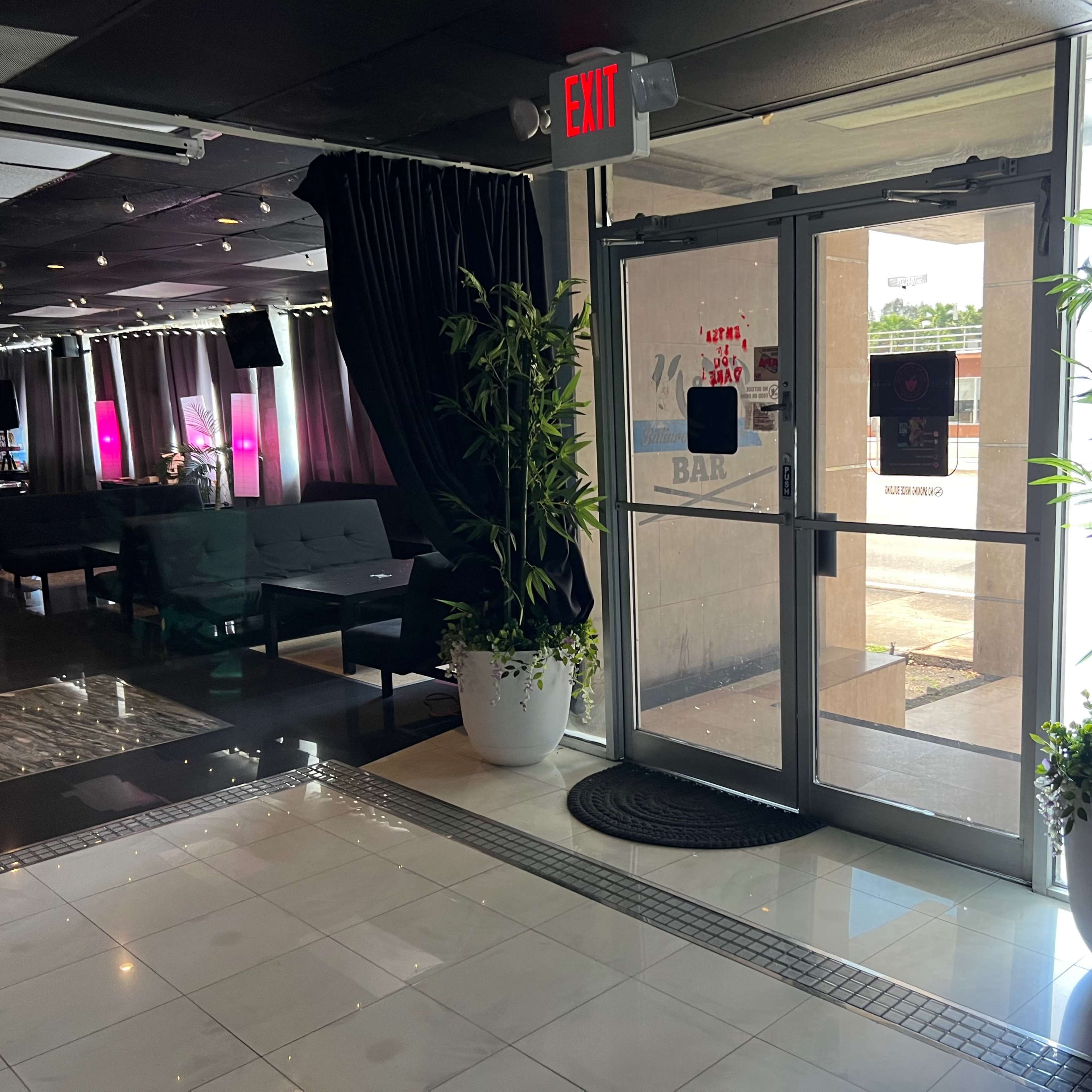 The interior of a lounge area featuring a glass entrance door, potted plants, and seating arrangements under ambient lighting.