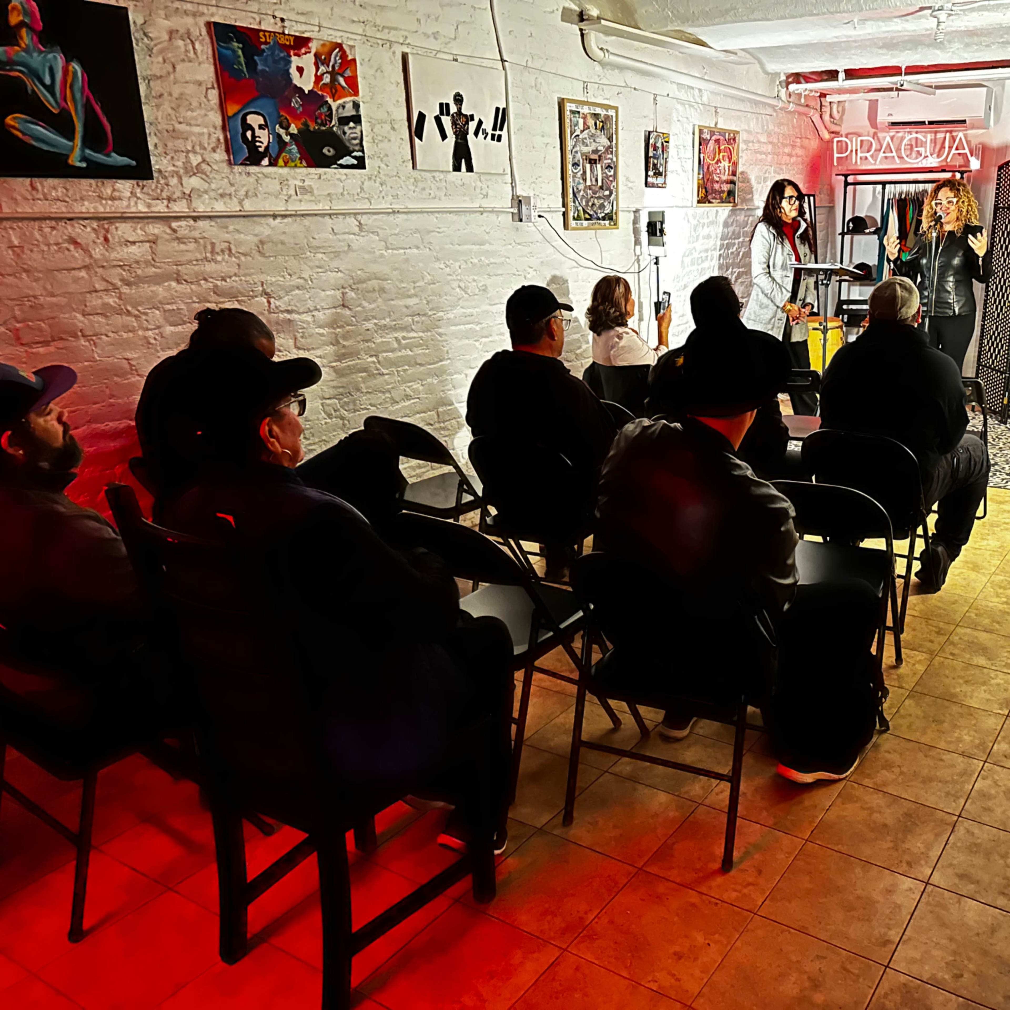 A small audience sits in rows of chairs as two performers speak in a dimly lit, art-adorned basement venue.