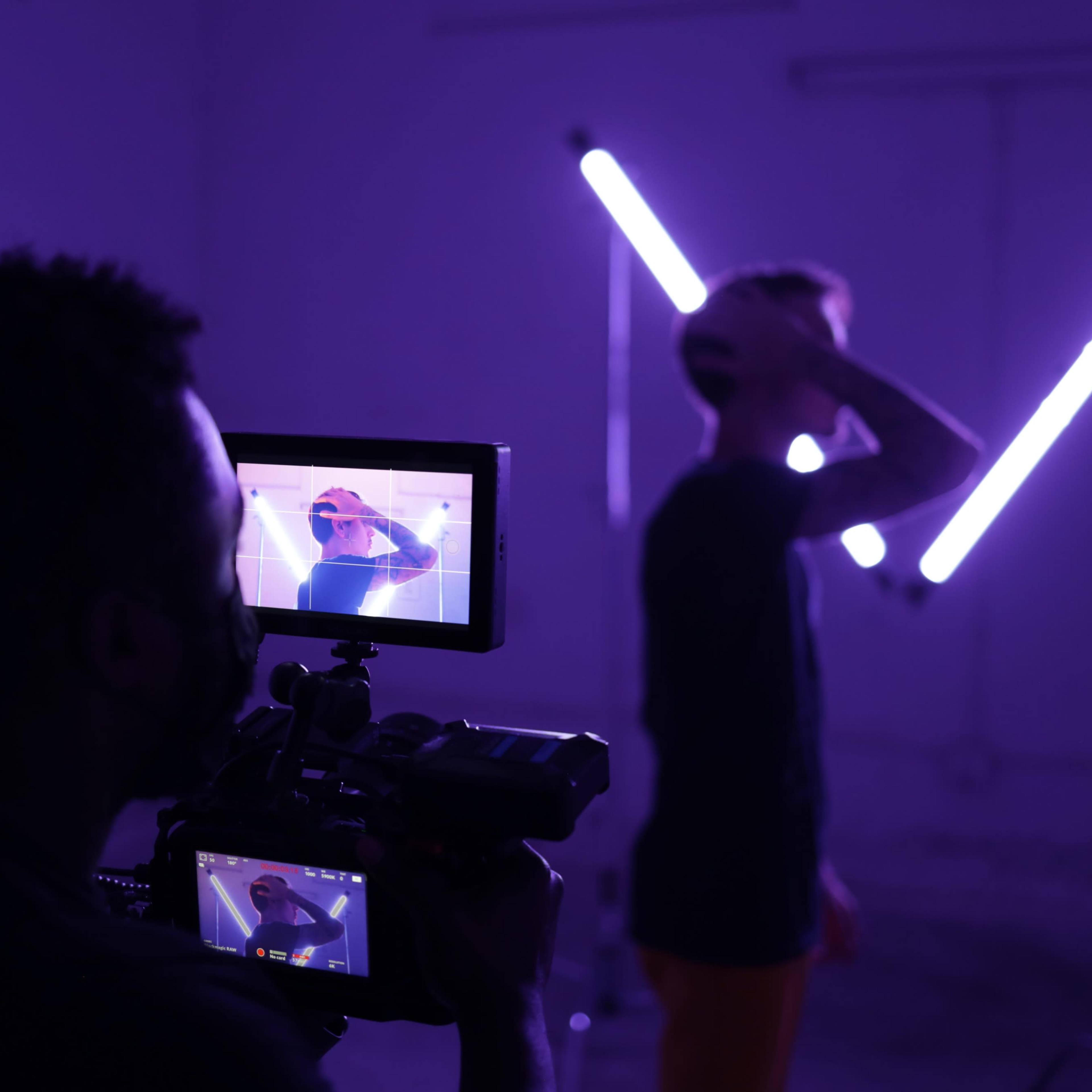 A person operates a camera while filming another individual against a backdrop of purple lighting.