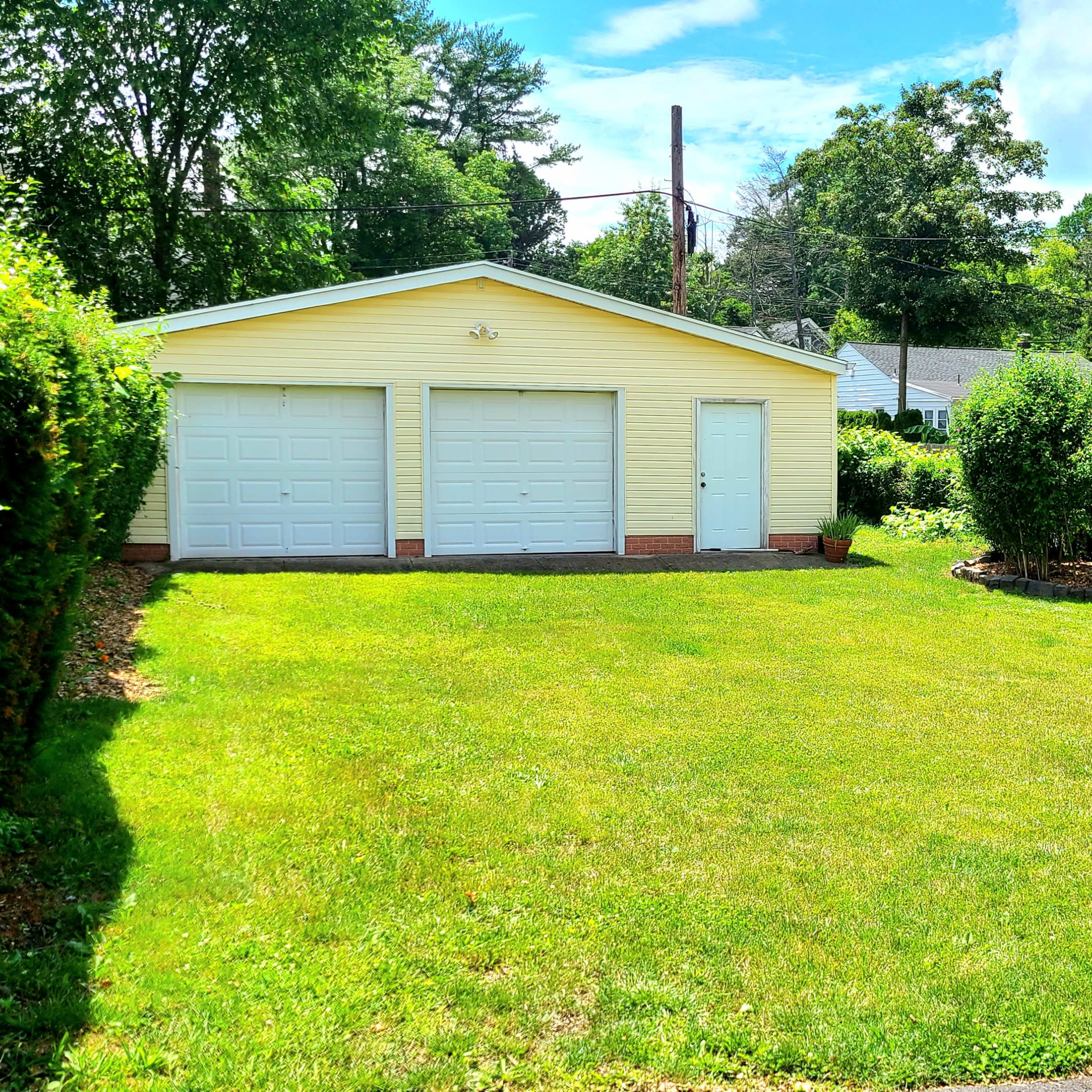 A two-car garage with a light yellow exterior is set in a grassy area surrounded by hedges, with additional buildings visible in the background.