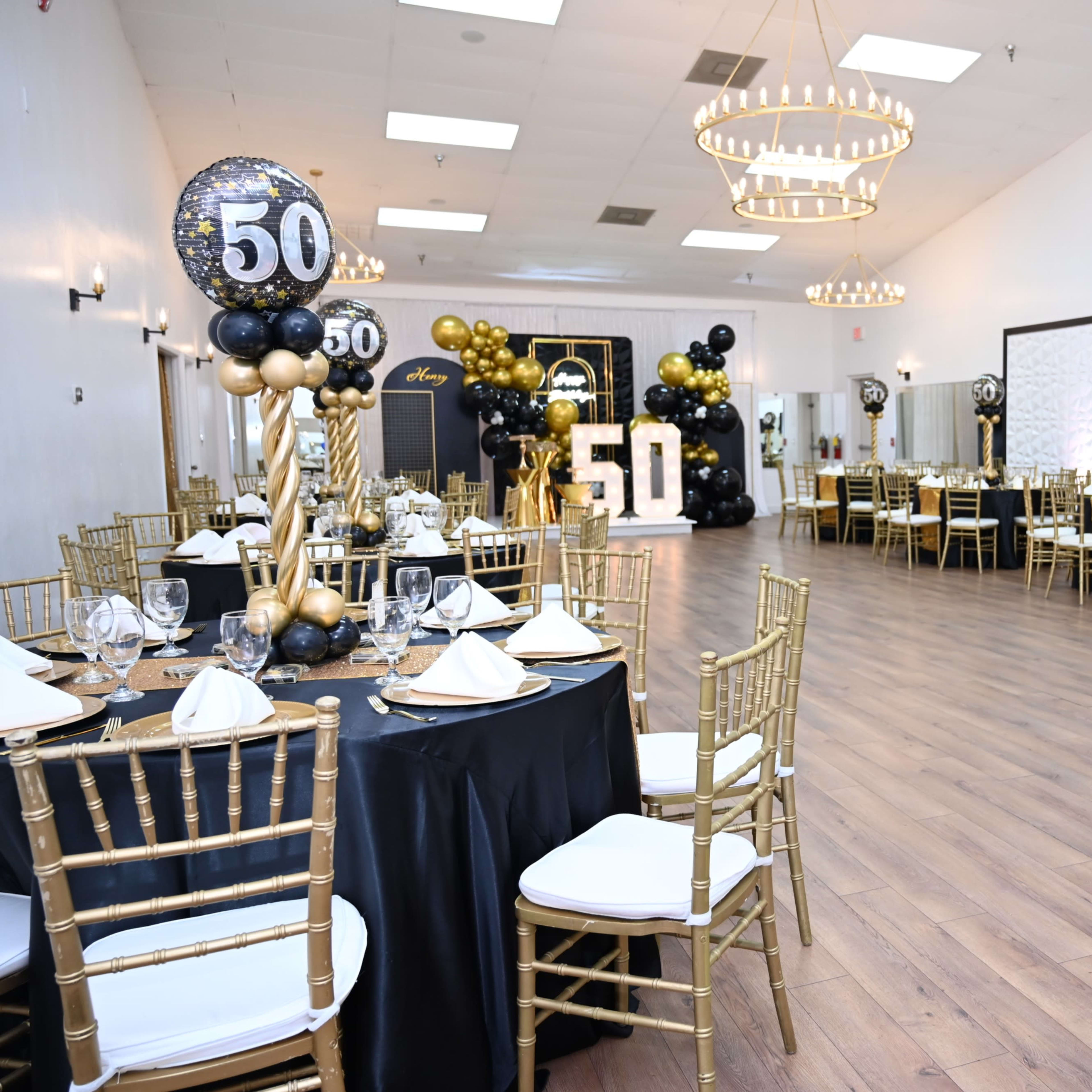 A spacious event hall decorated for a 50th birthday celebration, featuring black and gold table settings, elegant chairs, and decorative balloons.