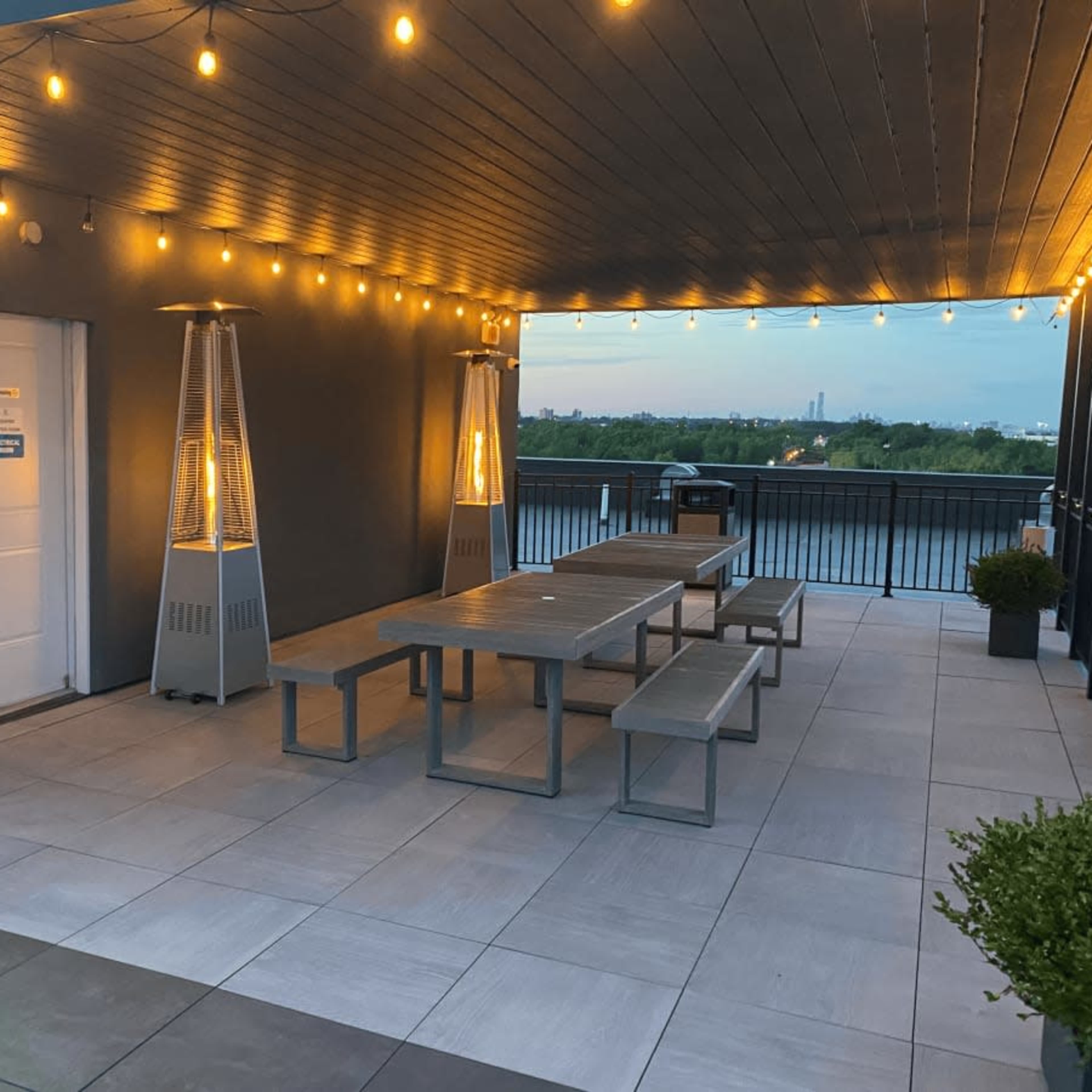 A rooftop area with several outdoor tables and benches, surrounded by string lights and two outdoor heaters, overlooking a cityscape.