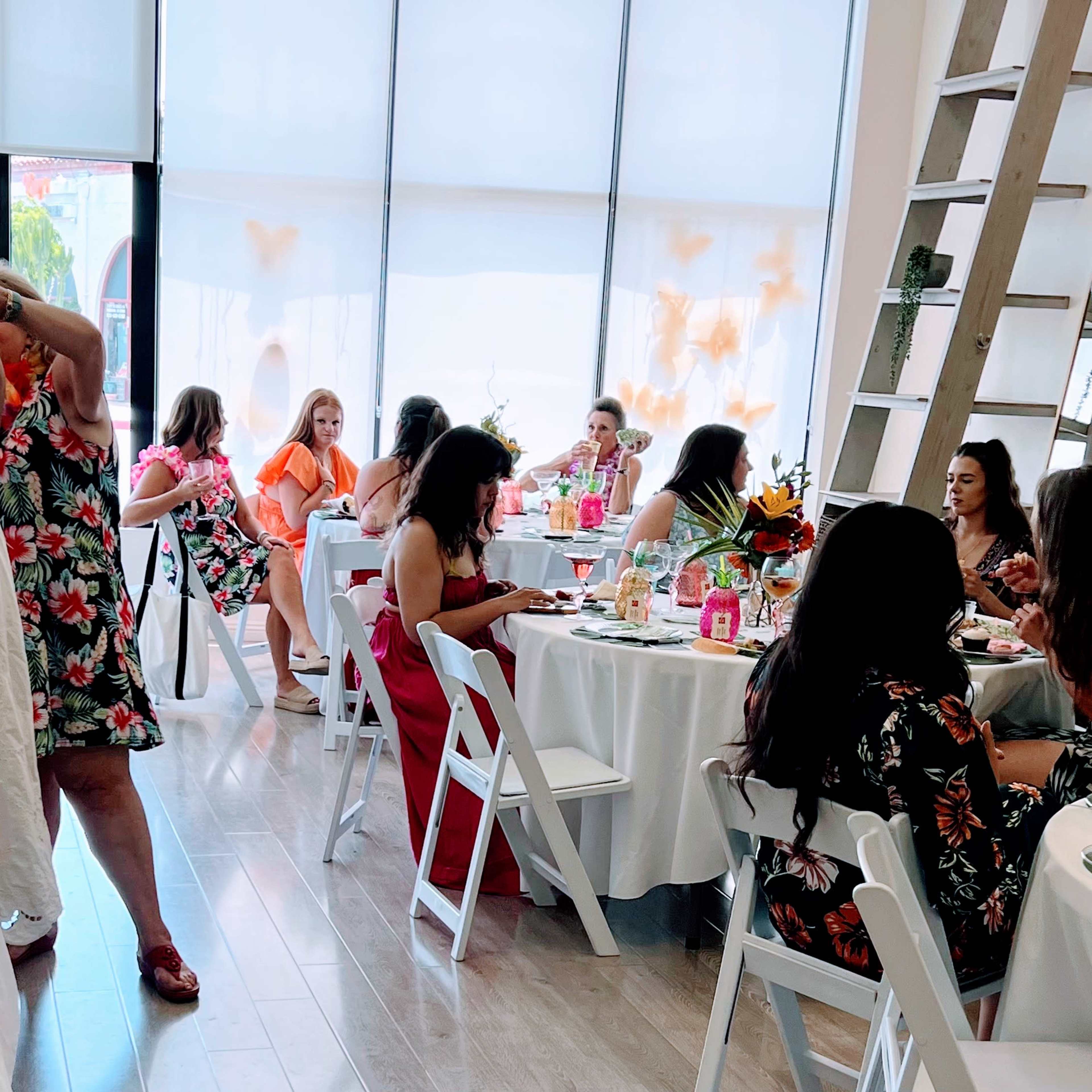 A group of women are seated at tables in a bright, airy room decorated with floral arrangements, enjoying food and drinks.