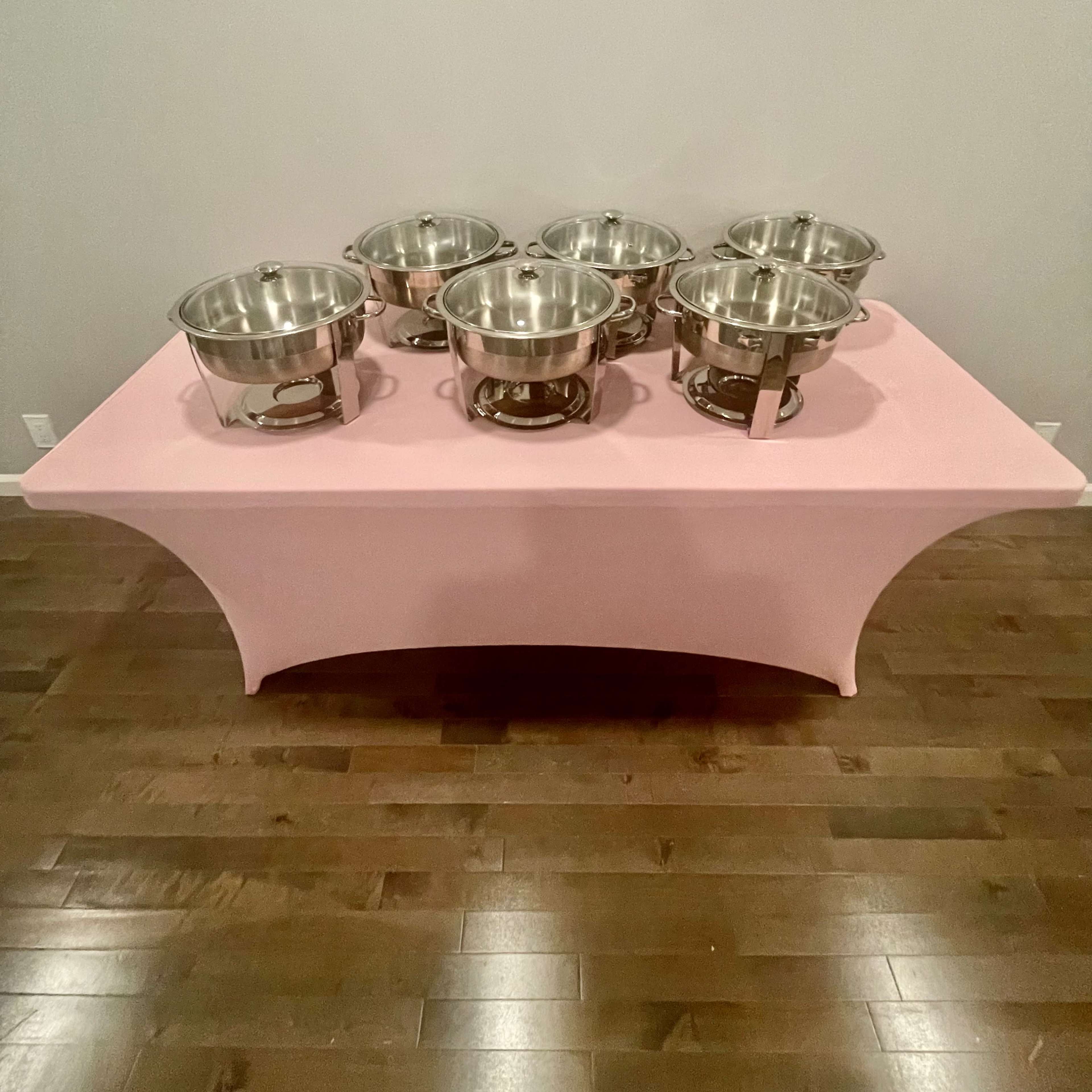 A table with a pink cover displays six stainless steel chafing dishes arranged in a row.