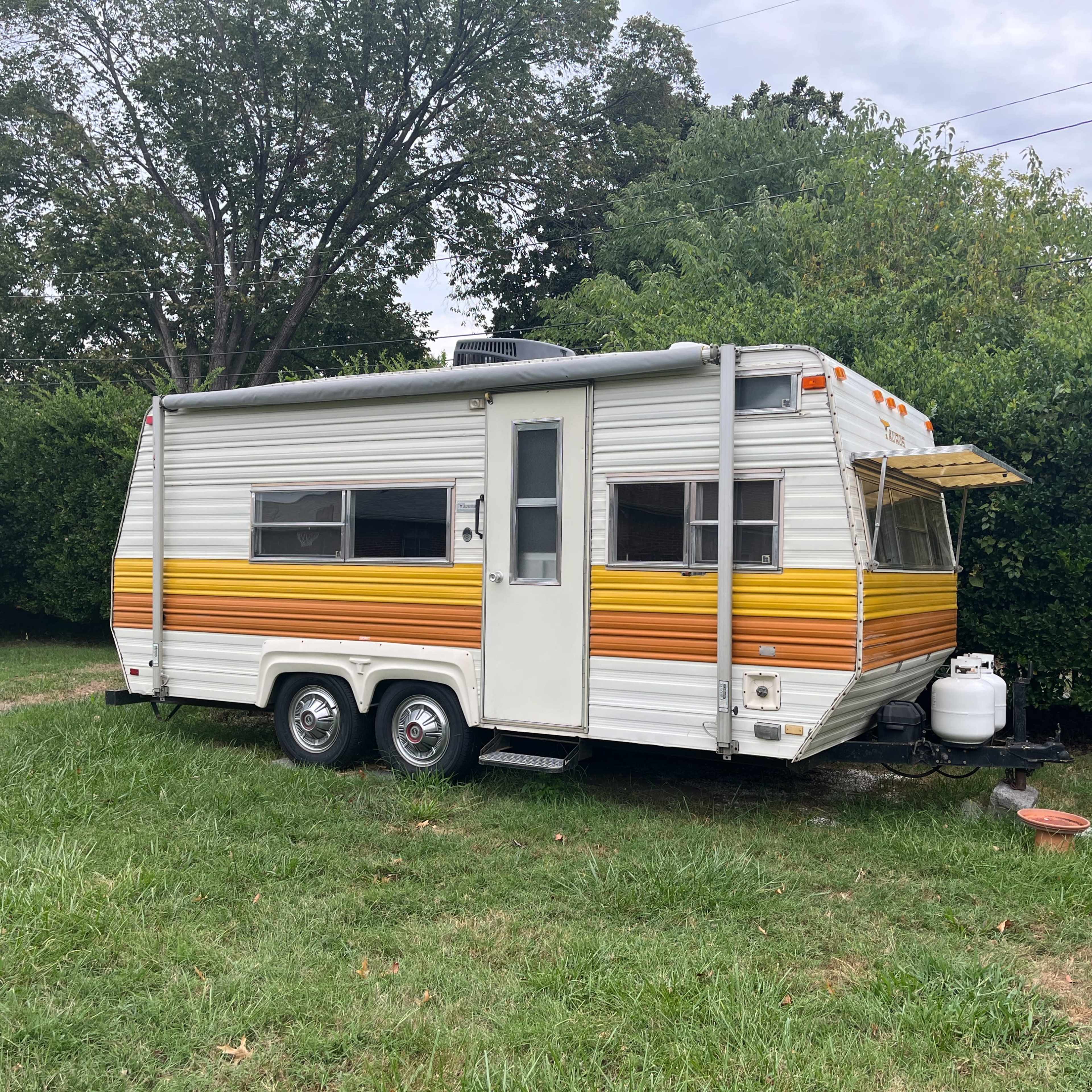 A vintage camper trailer with orange and yellow stripes is parked on grass next to a hedge.