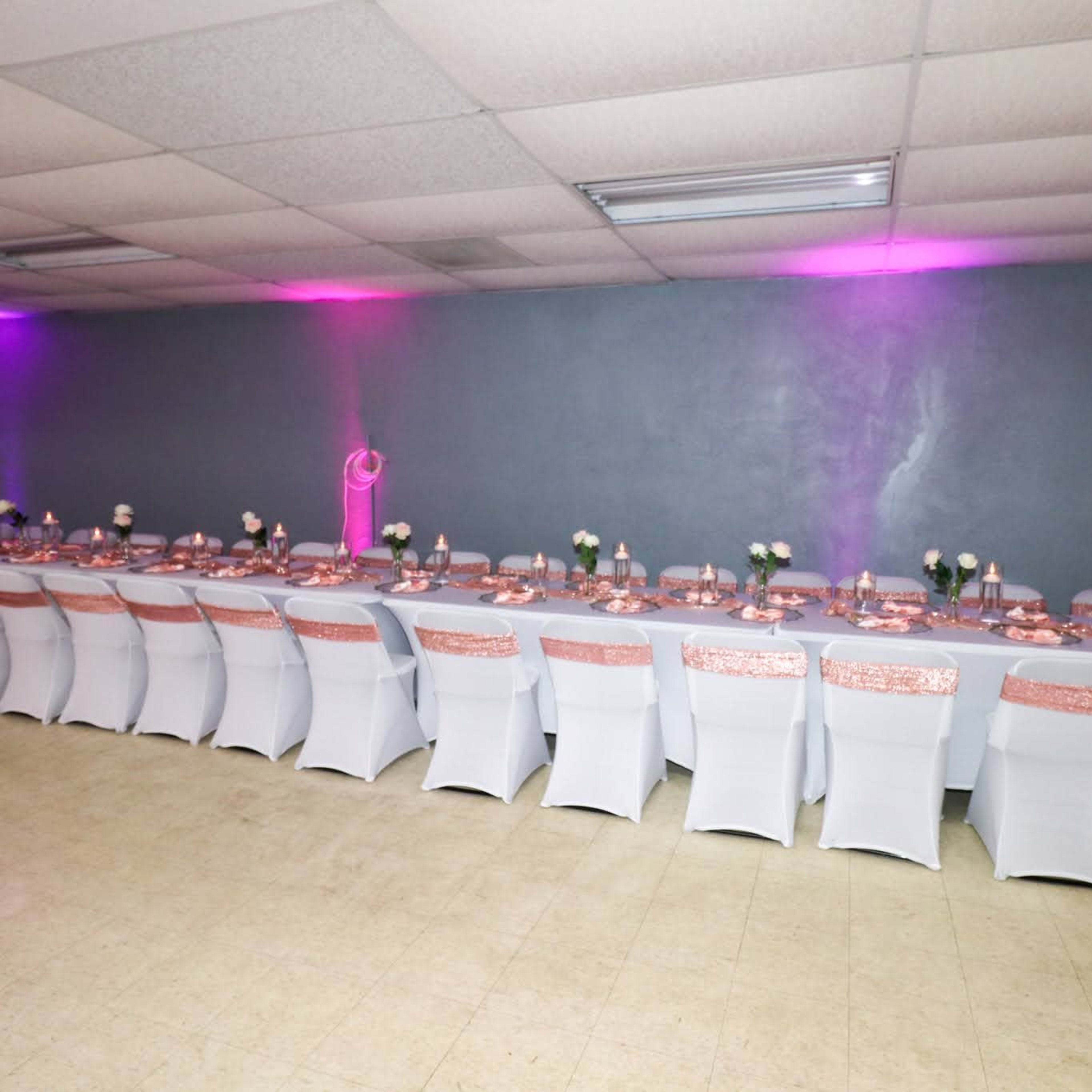A long banquet table is set with white tablecloths and rose gold accents in a dimly lit room with purple lighting.