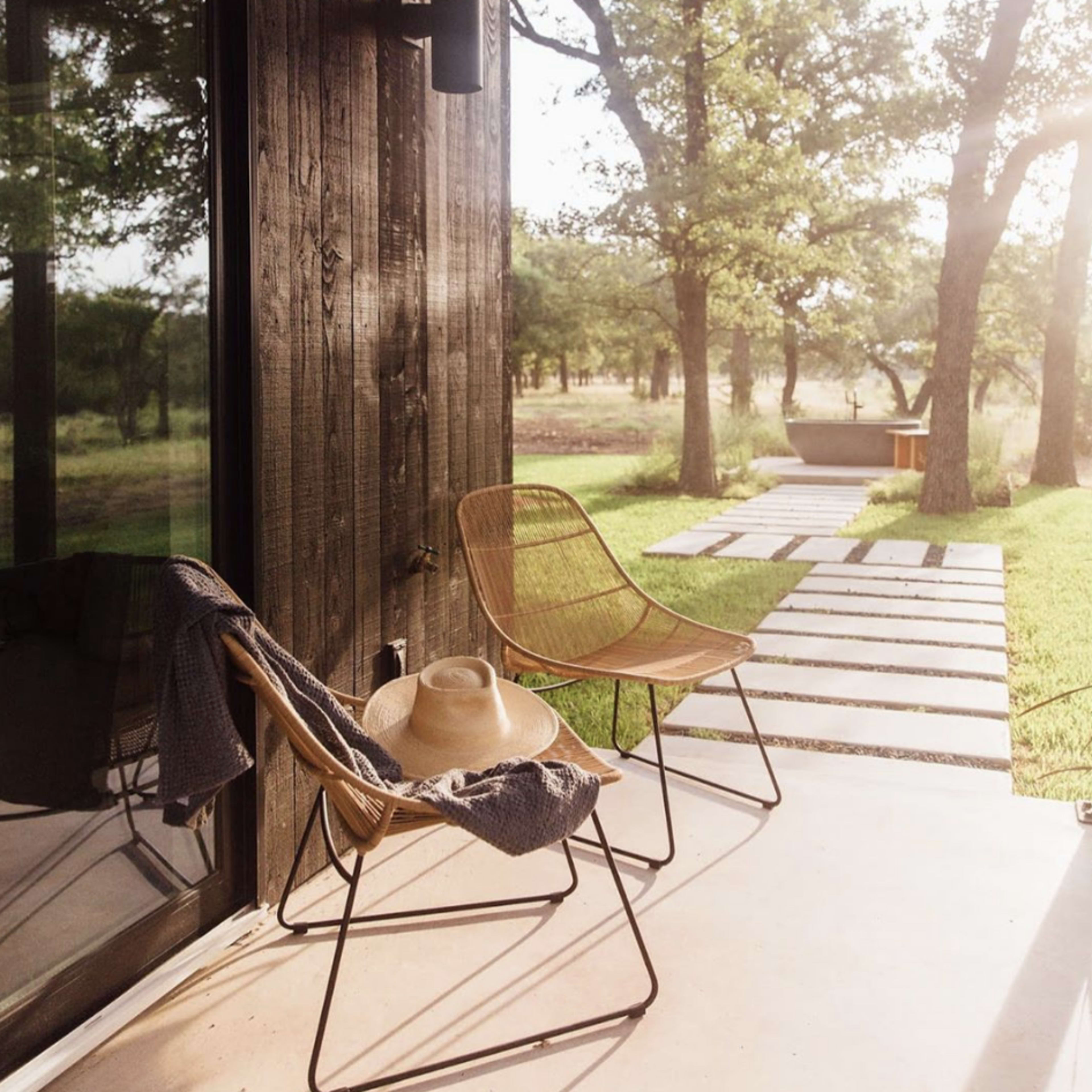 A porch with two chairs, a straw hat draped over one, and a pathway leading through a grassy area with trees in the background.