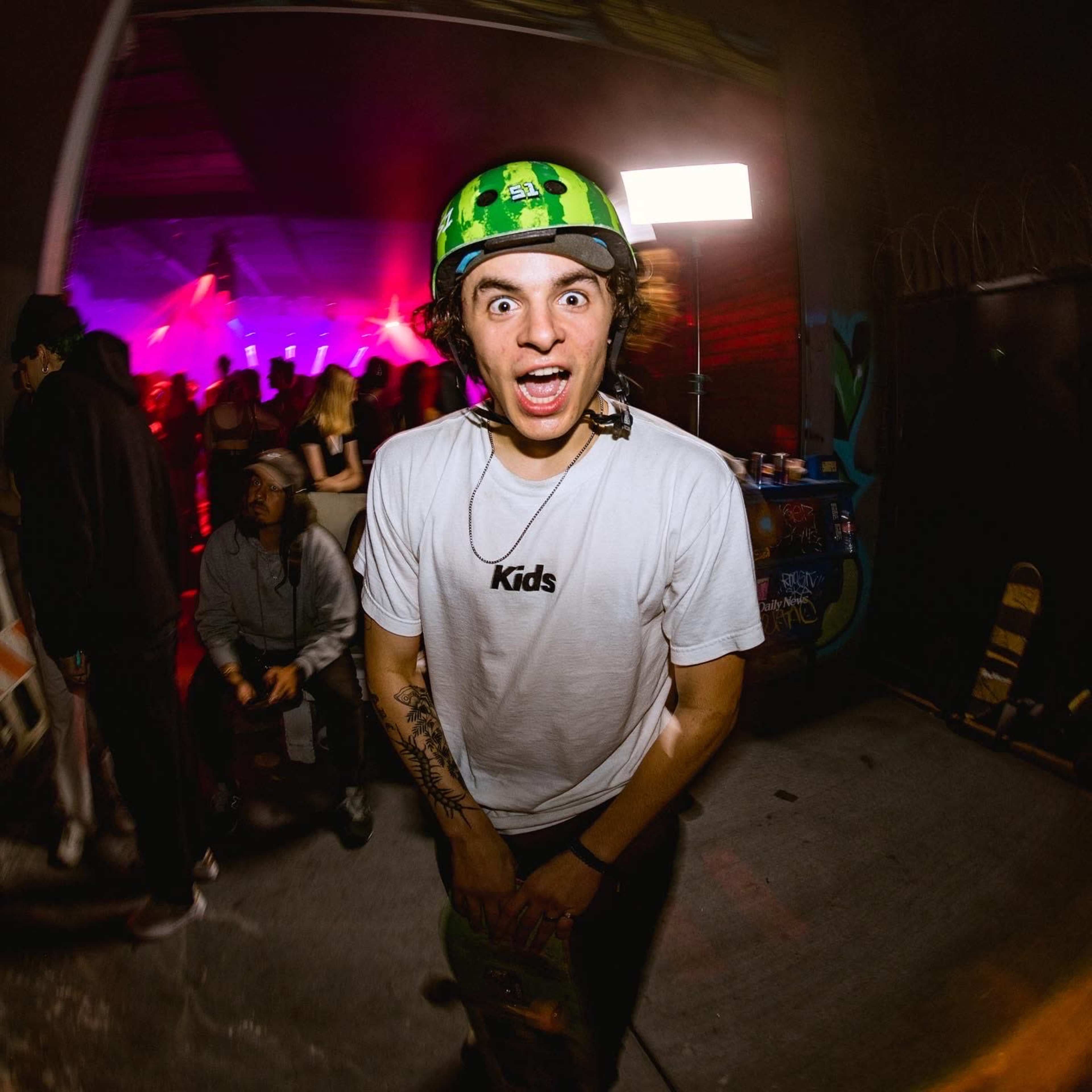 A young person wearing a green helmet and a "Kids" t-shirt stands with a skateboard in a vibrant, dimly lit setting filled with people.