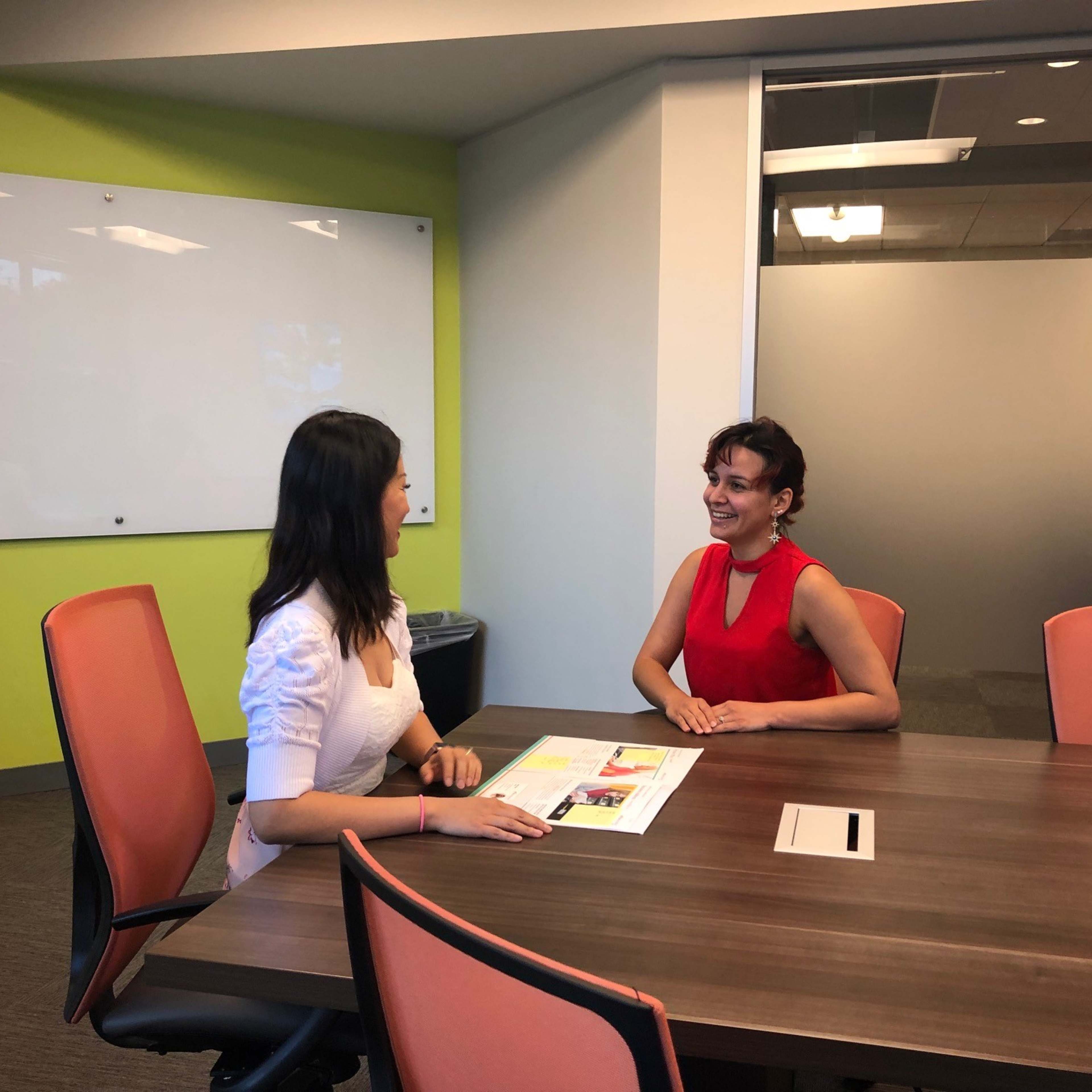 Two women are seated at a wooden conference table in a modern meeting room, discussing documents.