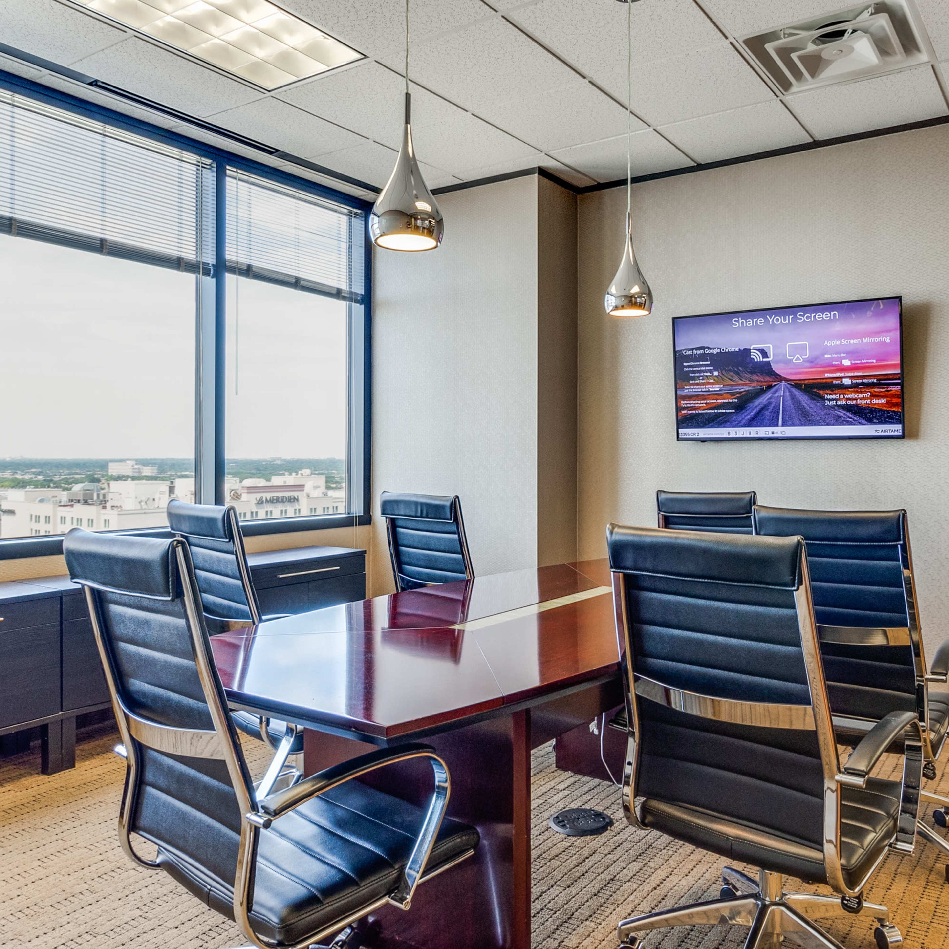 A conference room features a wooden table surrounded by black chairs, with a large window displaying a city view and a mounted screen displaying a digital presentation.