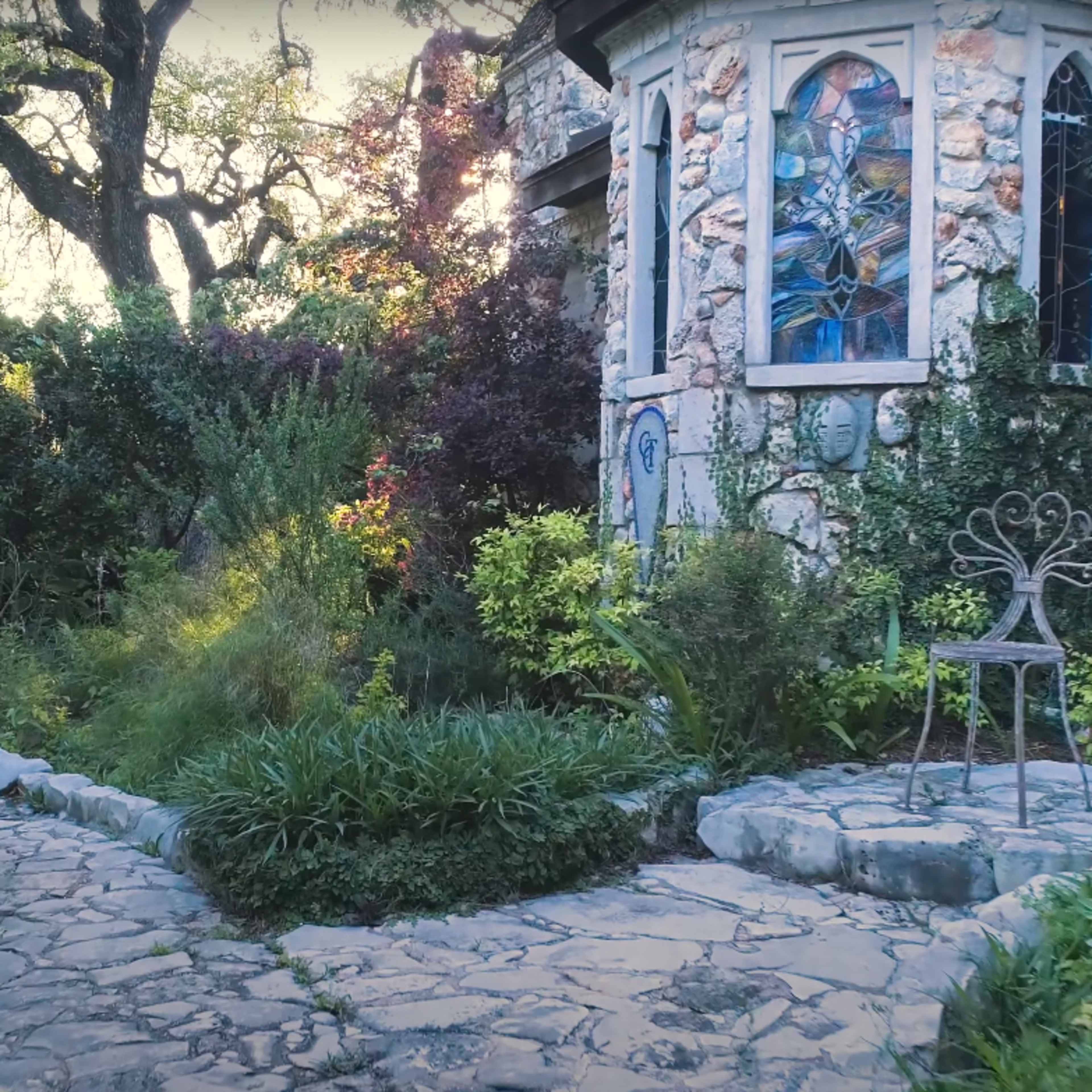 A stone pathway leading to a whimsical garden with lush greenery and a decorative structure featuring stained glass.