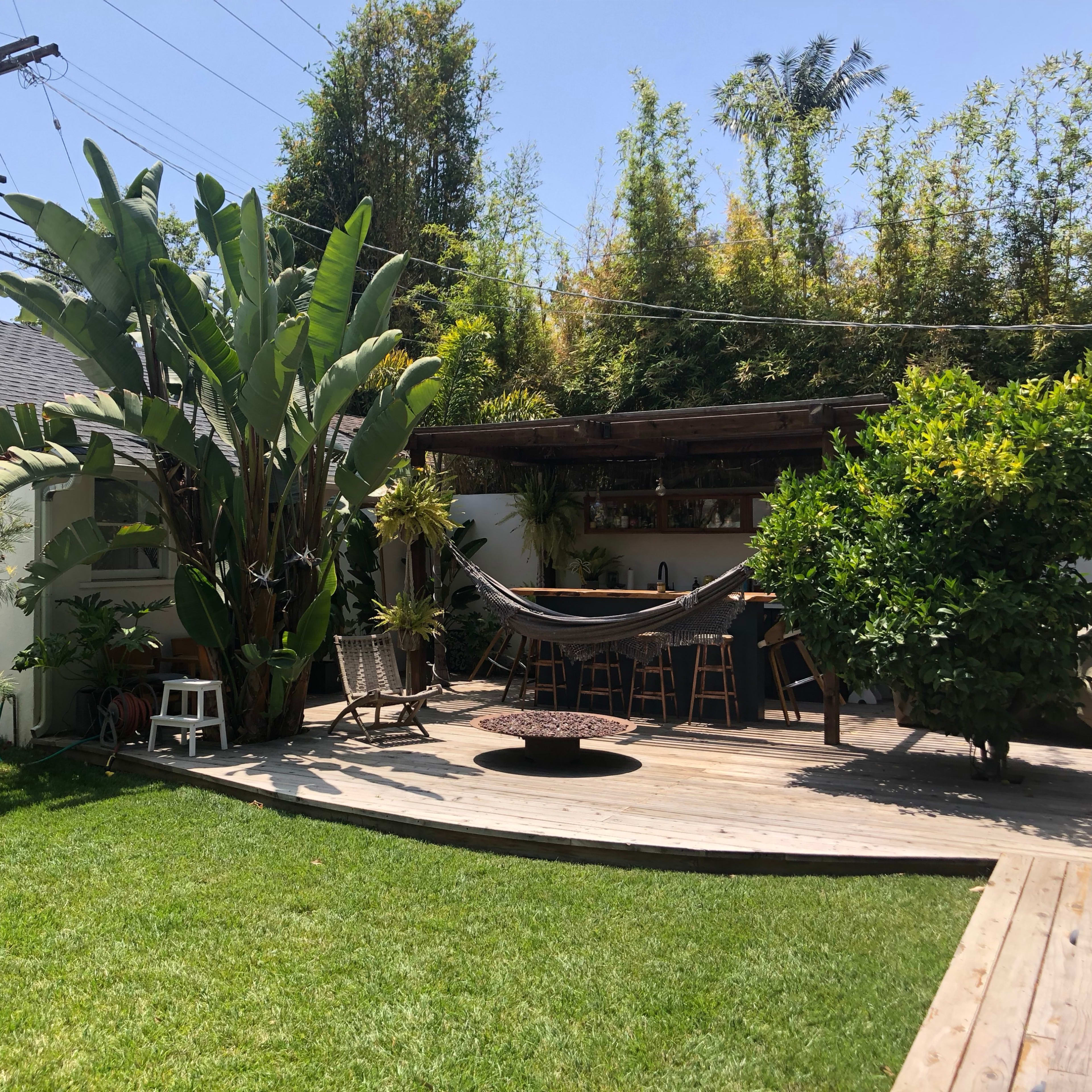 A dwelling with a wooden deck surrounded by lush greenery and large plants, featuring a hammock and outdoor dining area.