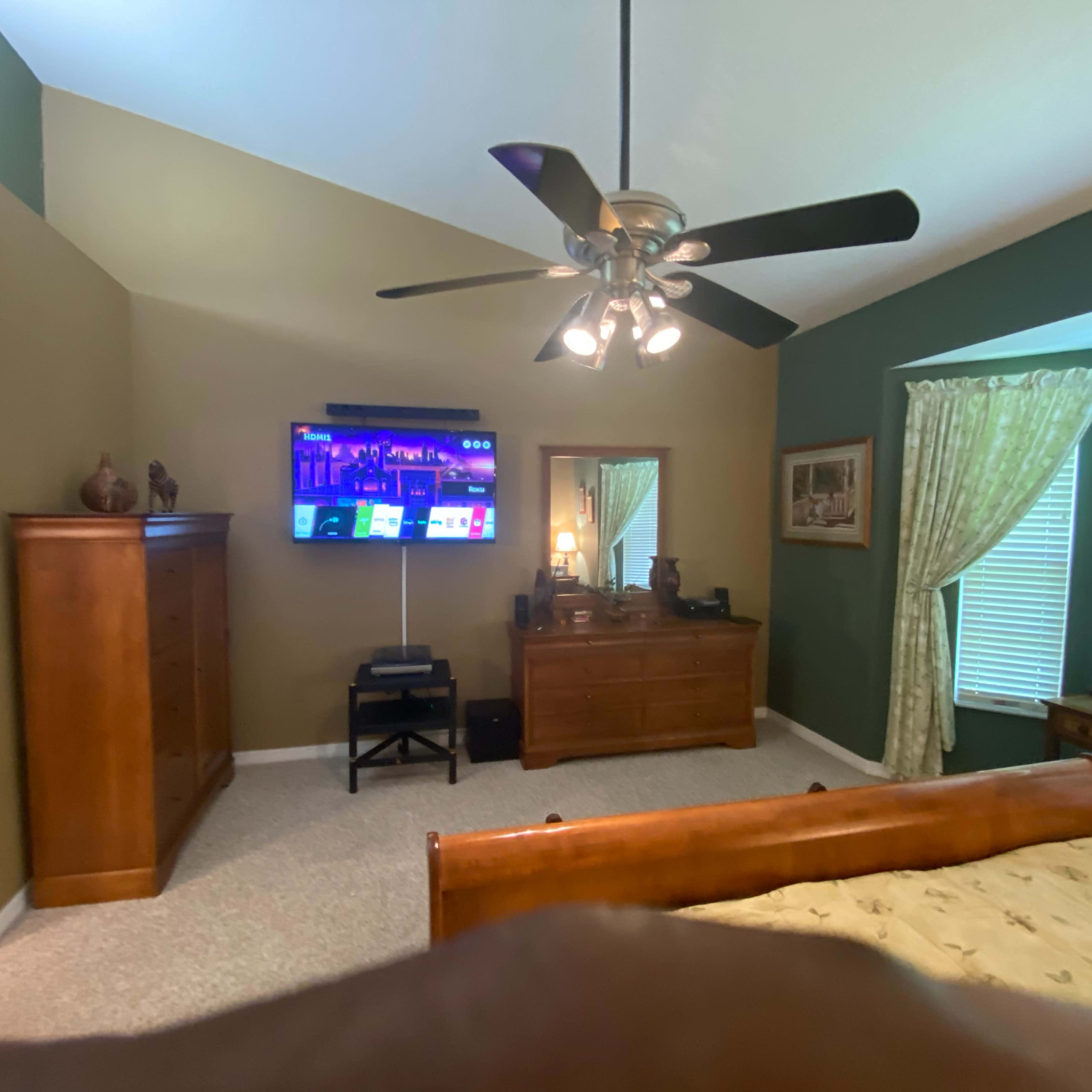 A bedroom features a wooden bed, a dresser, a television mounted on the wall, and windows with curtains, alongside an en-suite bathroom.