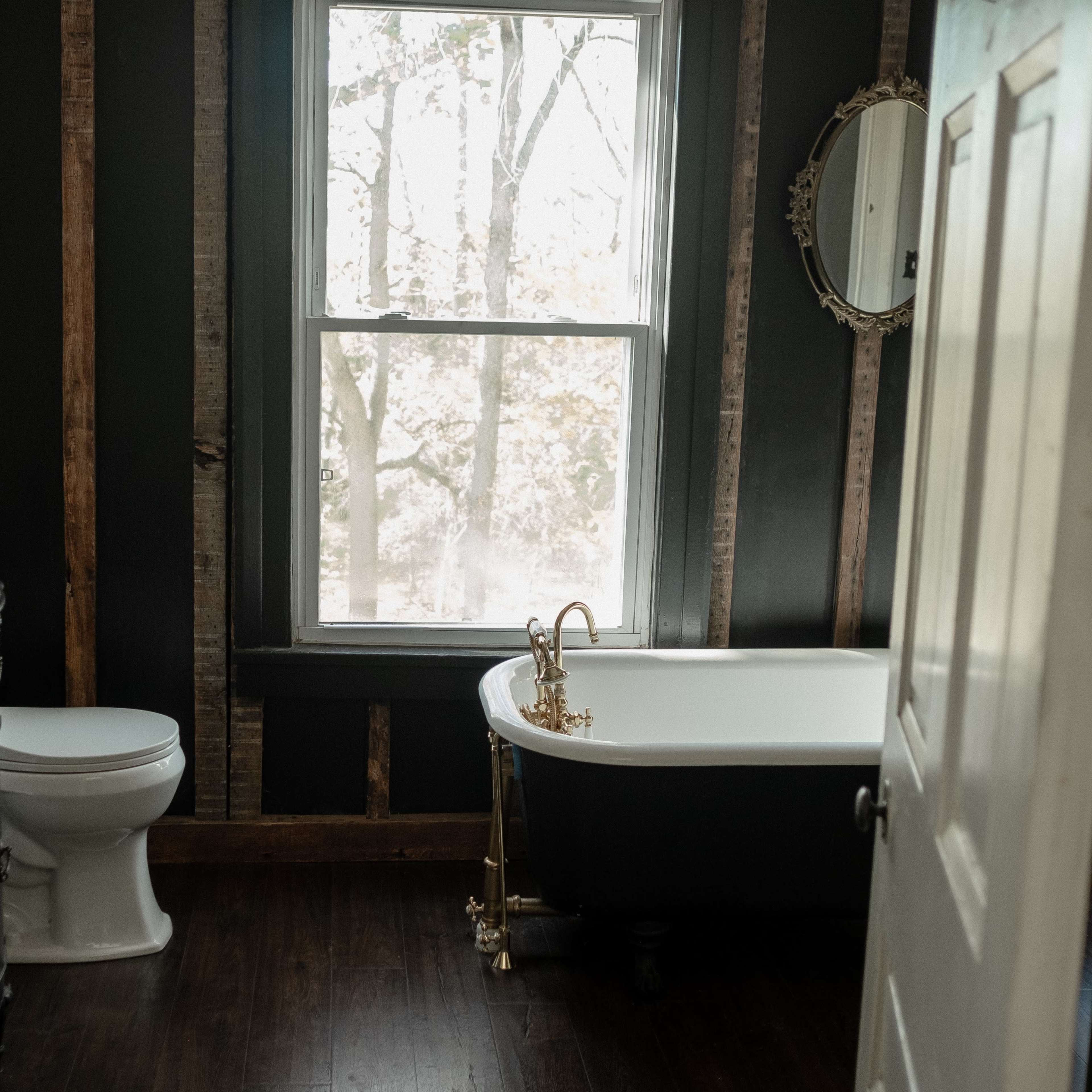 The bathroom features a freestanding tub, a white toilet, dual sinks with dark cabinetry, and a large window overlooking a forested area.
