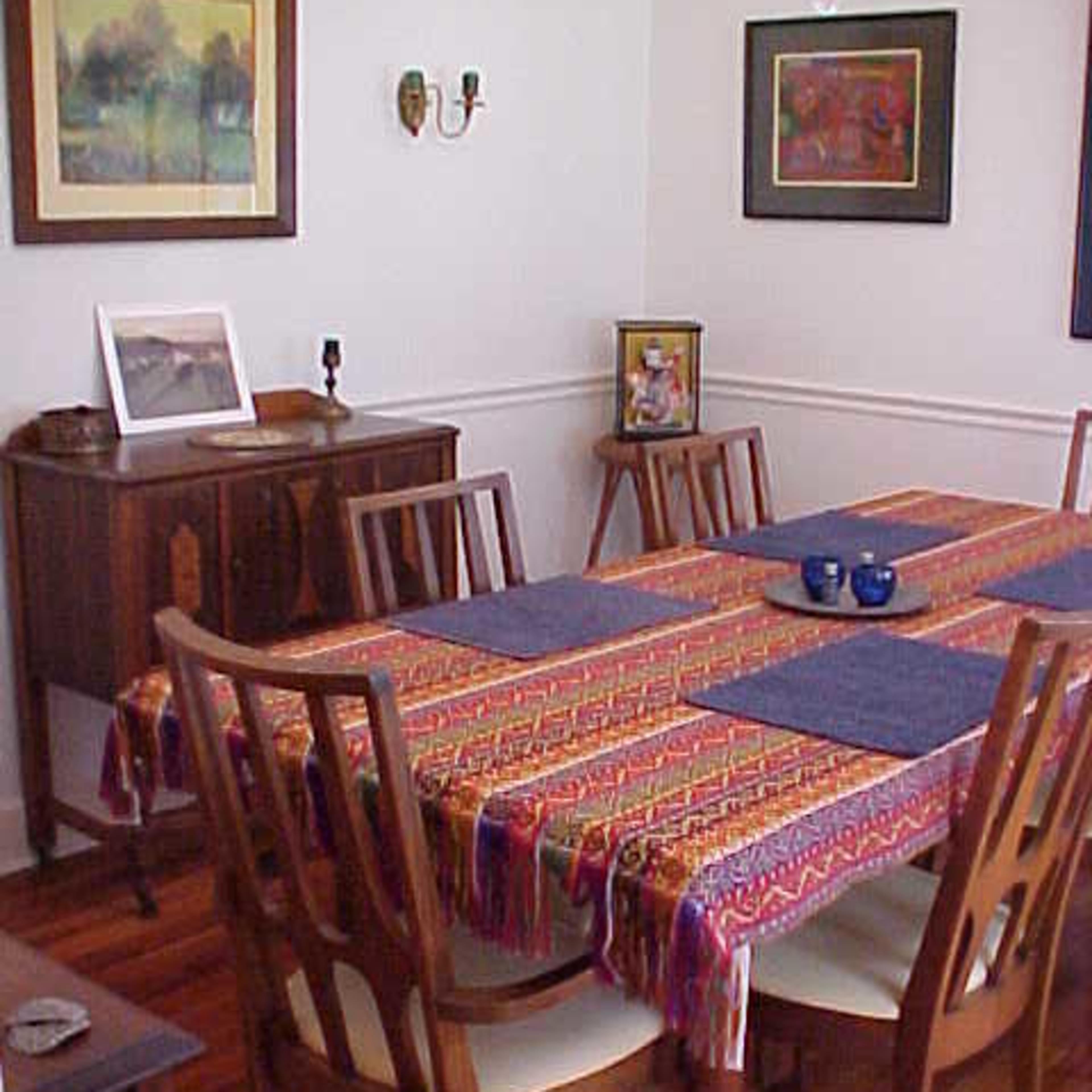 A dining room features a table set with a colorful tablecloth and surrounded by wooden chairs, with various framed artworks on the walls.