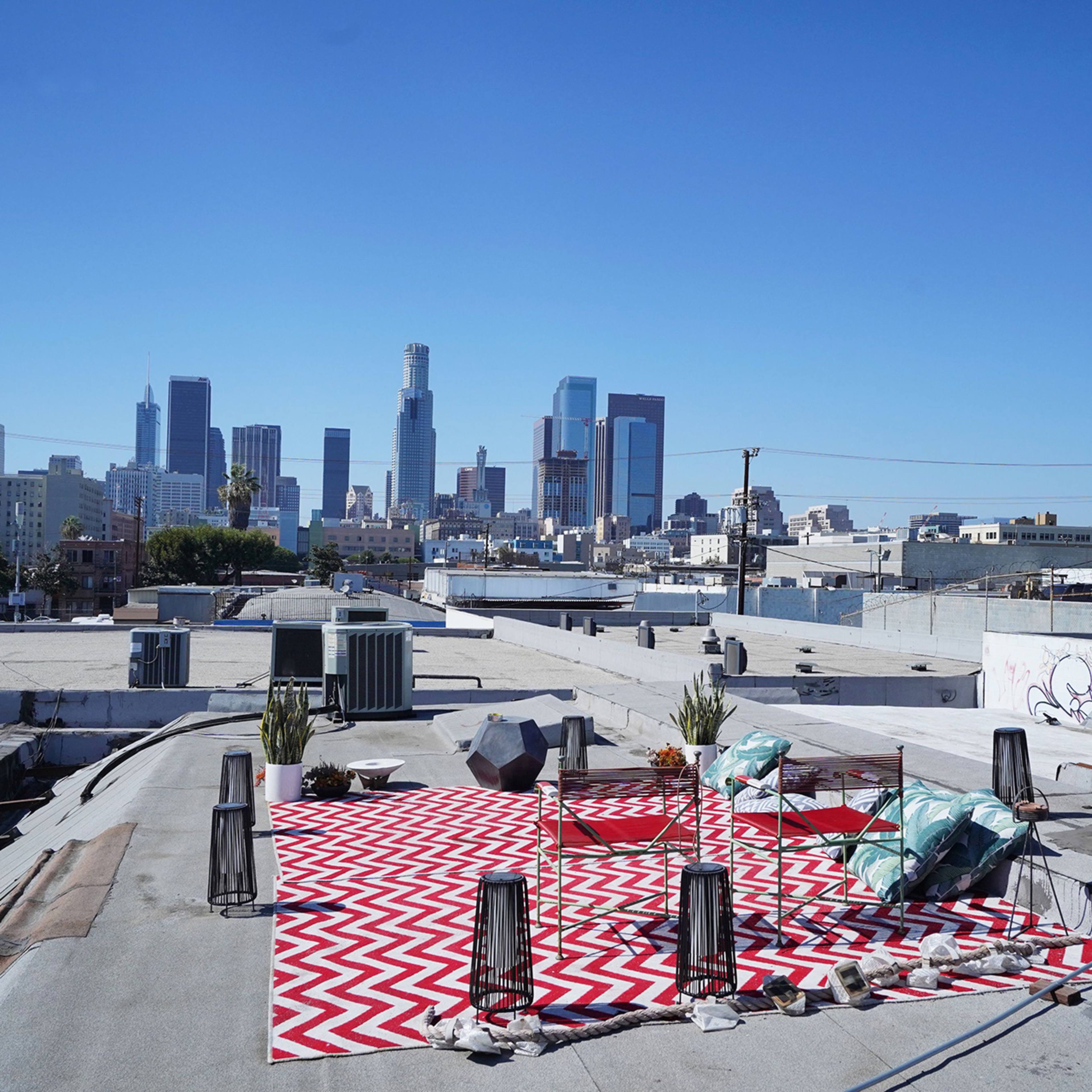 A rooftop terrace in an urban area, featuring a large zigzag-patterned rug, potted plants, and views of the Los Angeles skyline.