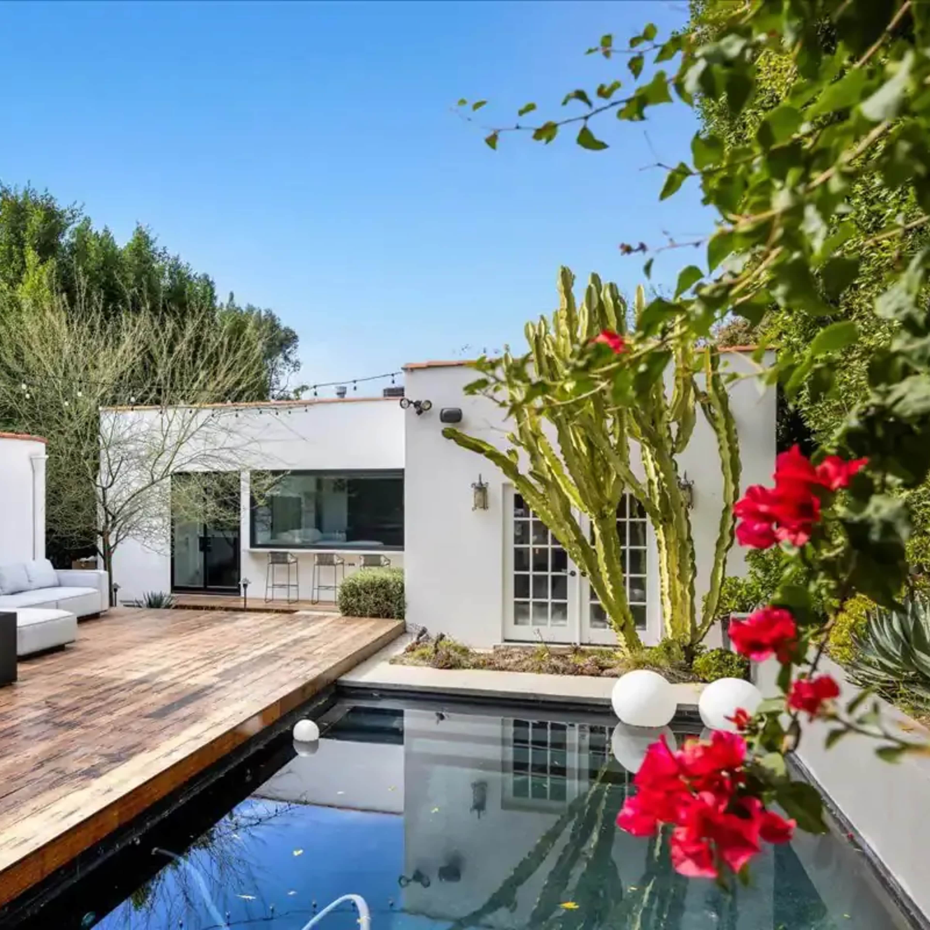 A modern backyard features a wooden deck with seating, a swimming pool, and surrounding greenery.